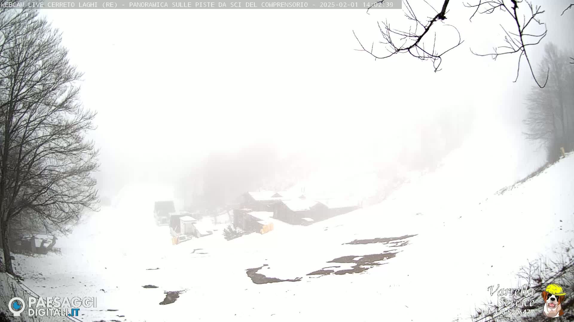 A snowy, foggy scene shows a few buildings nestled in a valley, surrounded by bare trees and snow-covered ground.