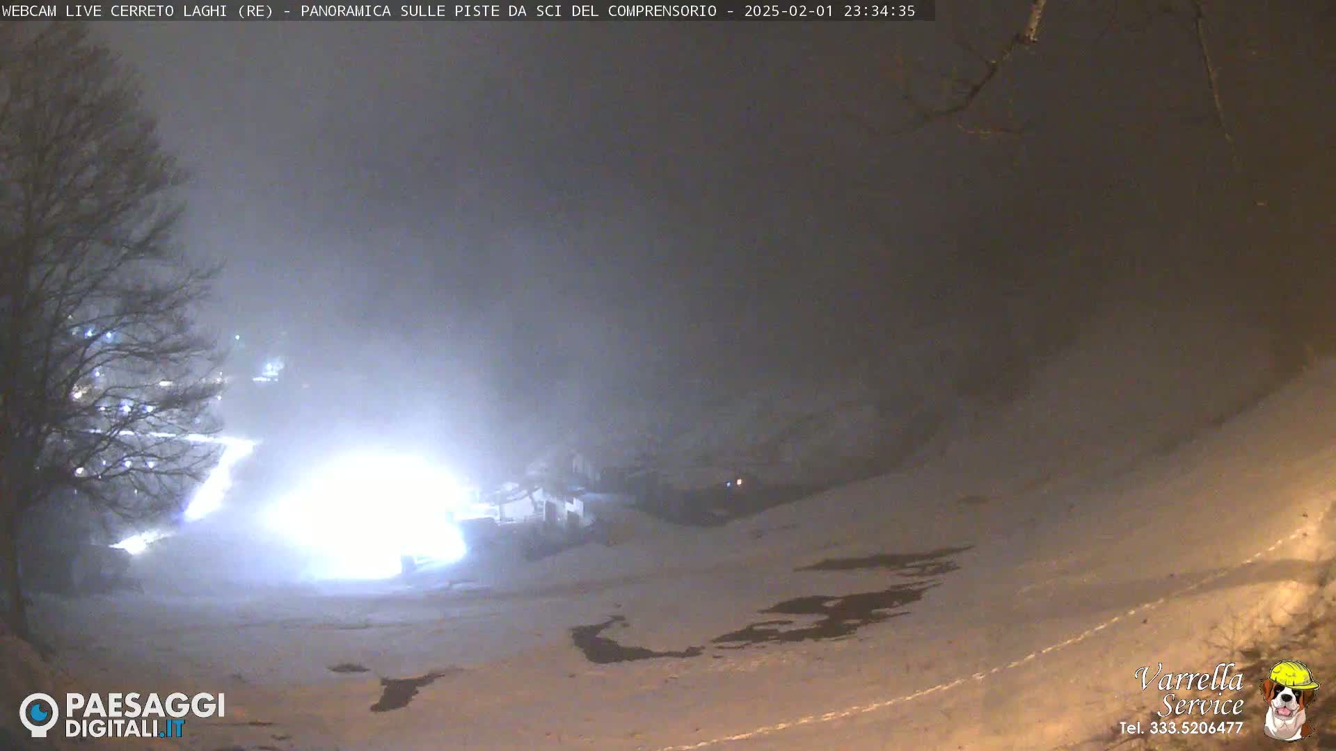 A snow-covered landscape at night is dimly lit by artificial light sources in foggy conditions.