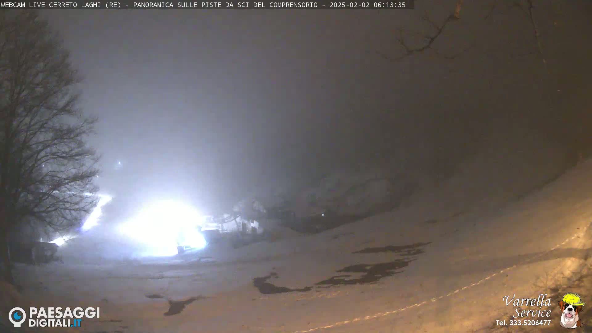 A snow-covered landscape is dimly lit at night under foggy conditions, with bright lights visible in the middle distance.