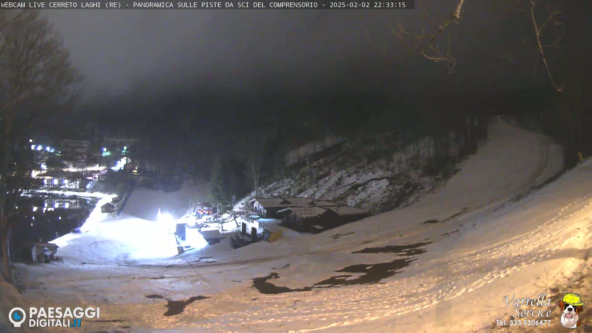 A nighttime view of a snow-covered ski slope with several buildings at the bottom, under a dark, hazy sky.