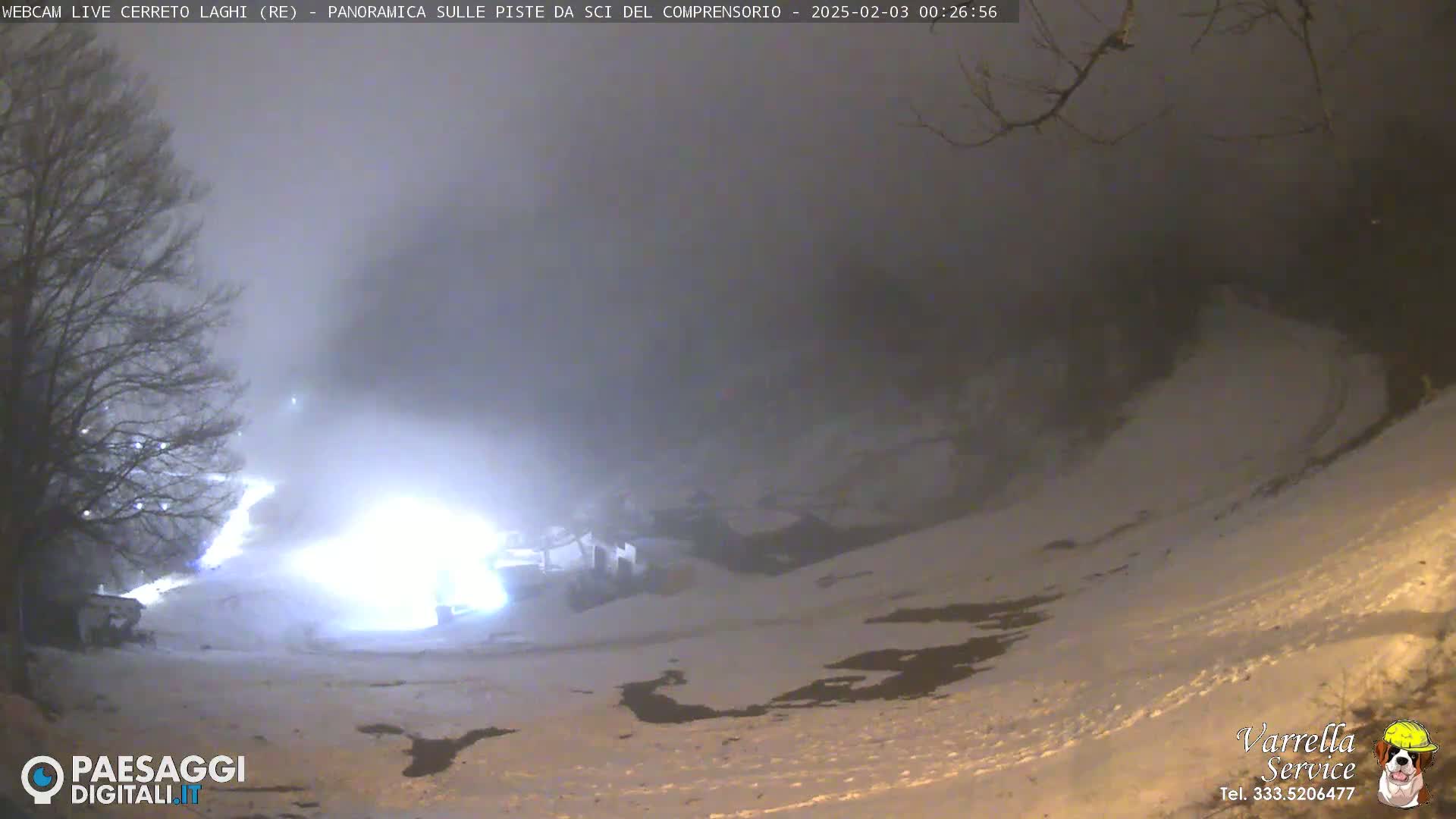 A snowy, foggy nighttime scene shows a brightly lit area at the bottom of a gently sloping hill.