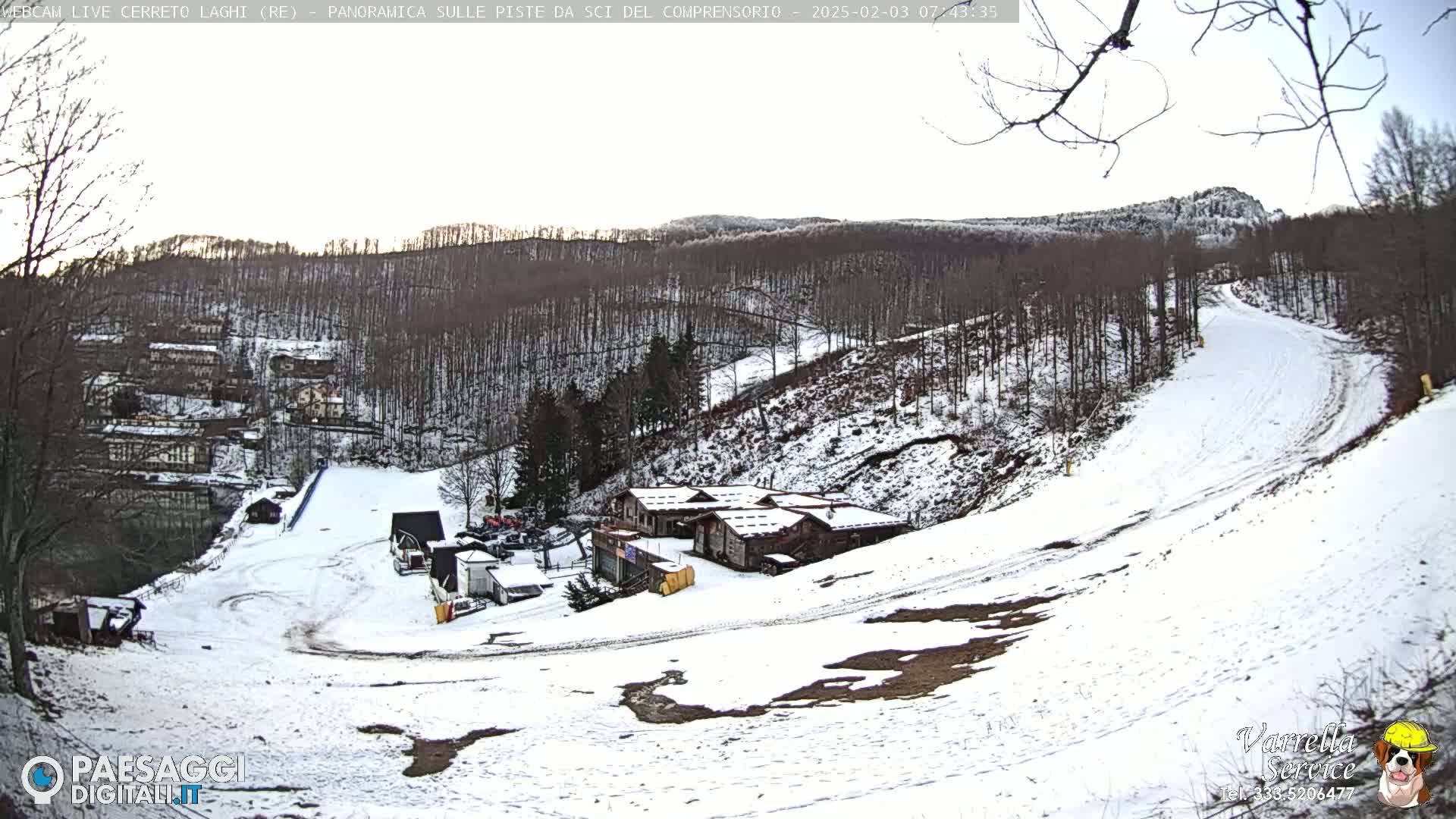 A snow-covered ski slope winds through a wooded hillside, with several buildings visible near the bottom of the slope under a mostly clear sky.