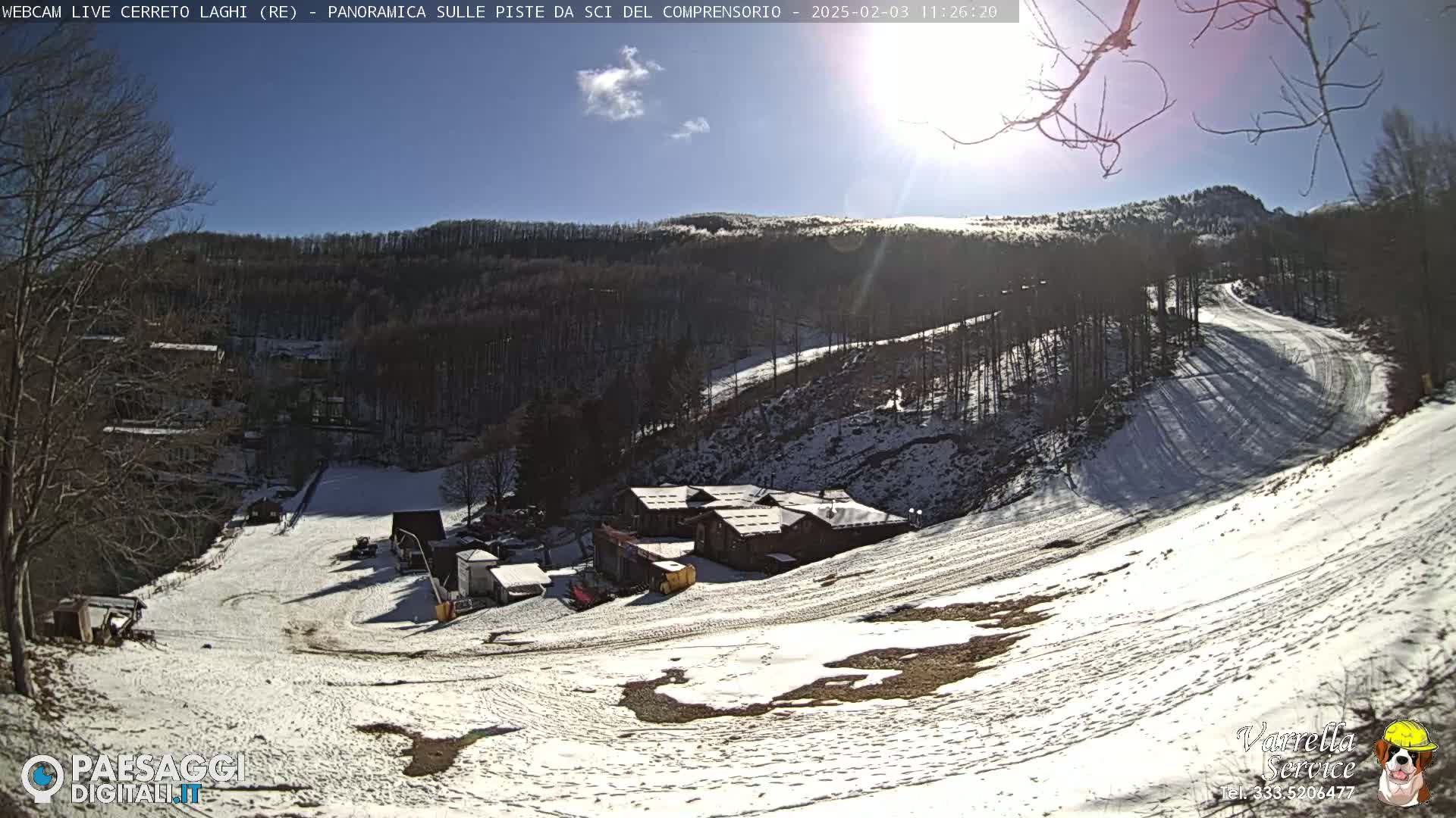 A sunny, snow-covered ski slope with a building and several trees in the background.