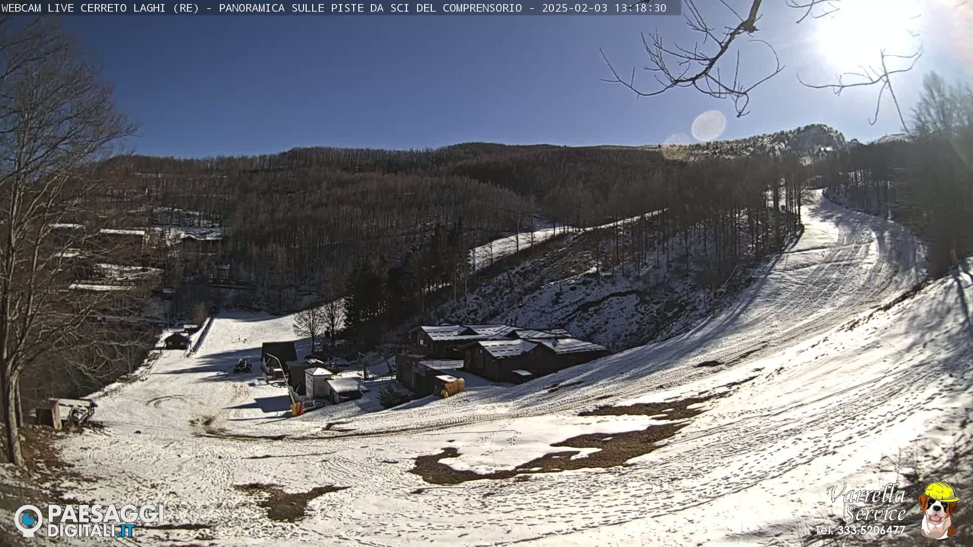 A sunny, snow-covered ski slope with a few buildings and bare trees in the background.