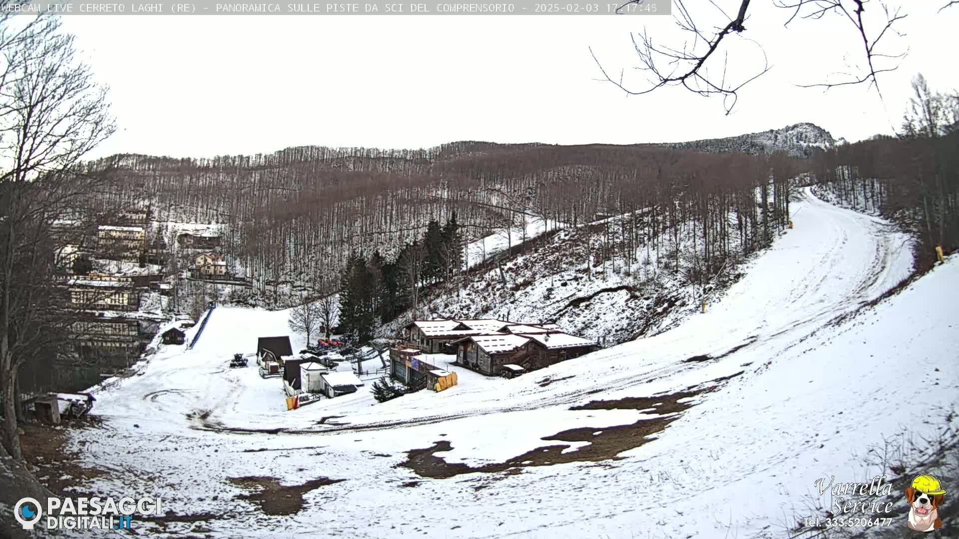 A snow-covered ski slope winds down a hillside through a sparsely wooded area, with several buildings visible near the bottom of the slope.