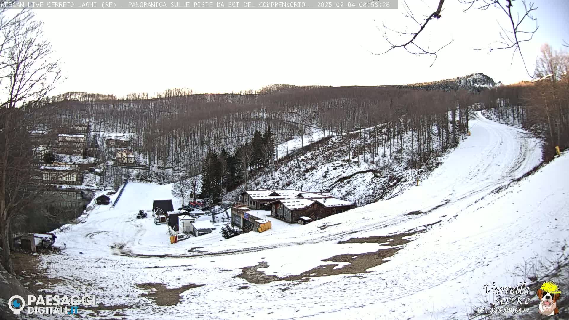 A snow-covered ski slope winds down a hill through a bare, snow-dusted forest to a cluster of buildings.
