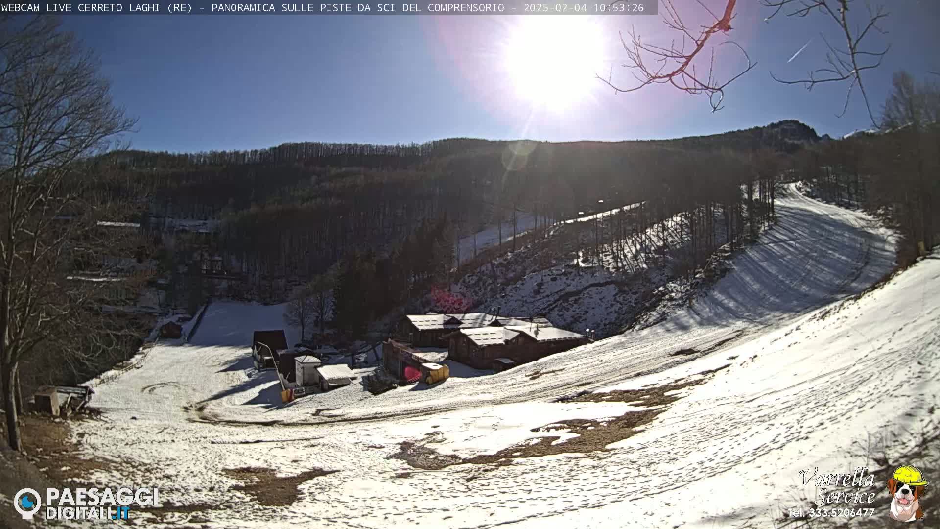 A sunny, snow-covered ski slope winds down a hillside through a sparsely wooded area, with a cluster of buildings near the bottom.