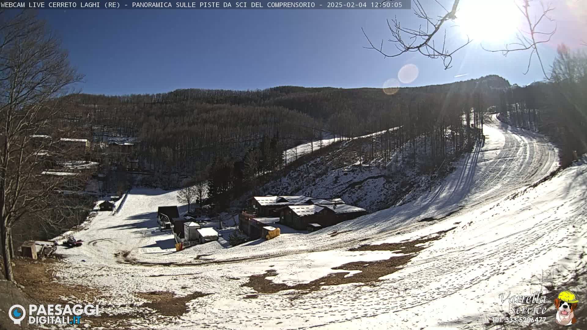 A sunny day at a snow-covered ski resort shows several ski runs, a cluster of buildings, and some sparse trees.