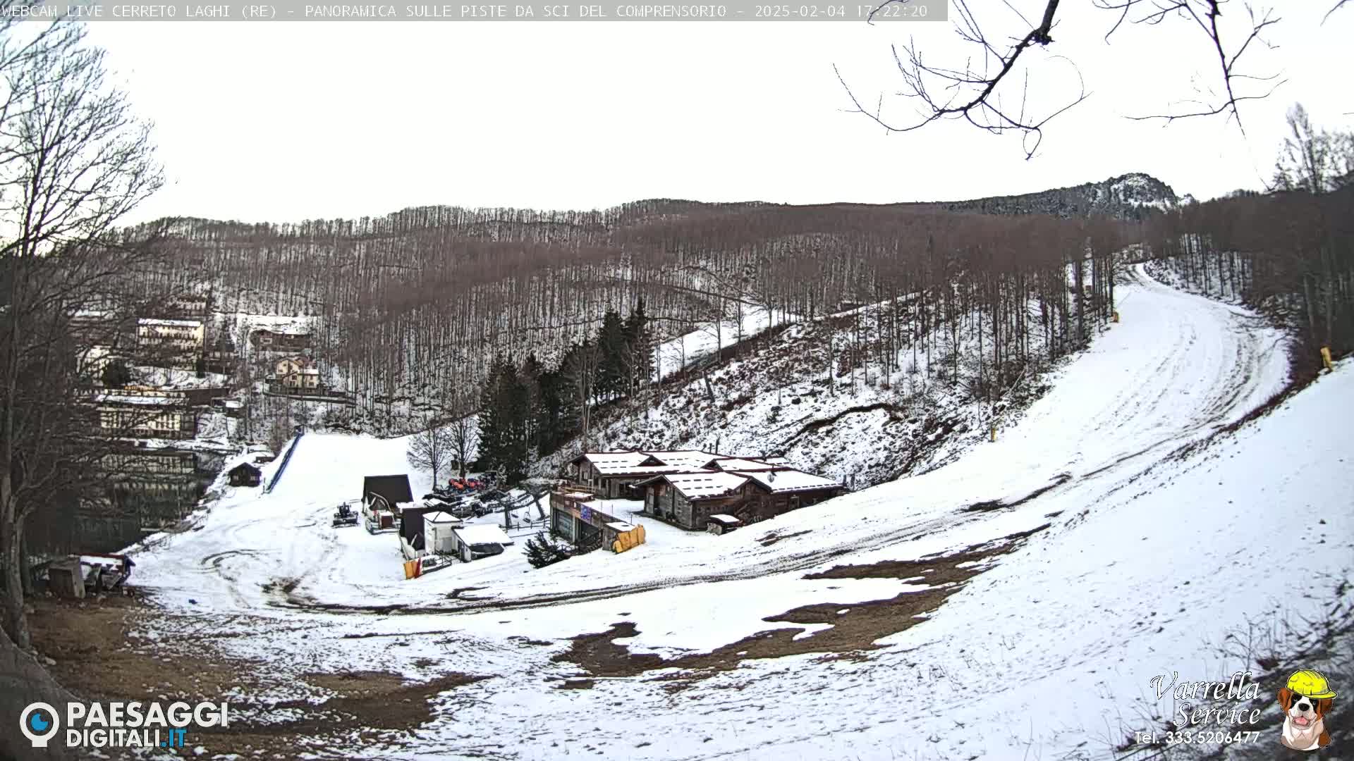 A snow-covered ski slope winds through a sparsely wooded hillside, with a cluster of buildings at the bottom of the slope under an overcast sky.