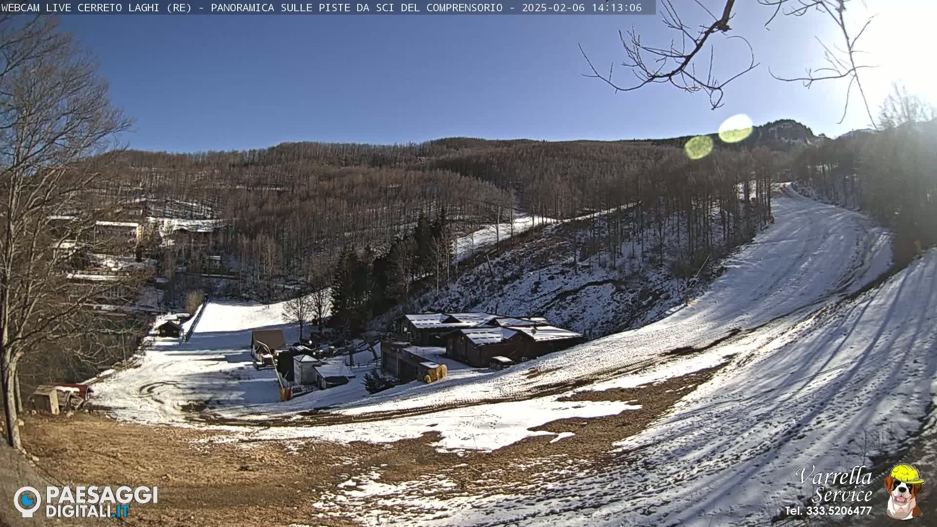 A sunny, snow-dusted ski slope descends into a valley with a cluster of buildings and bare trees under a clear blue sky.