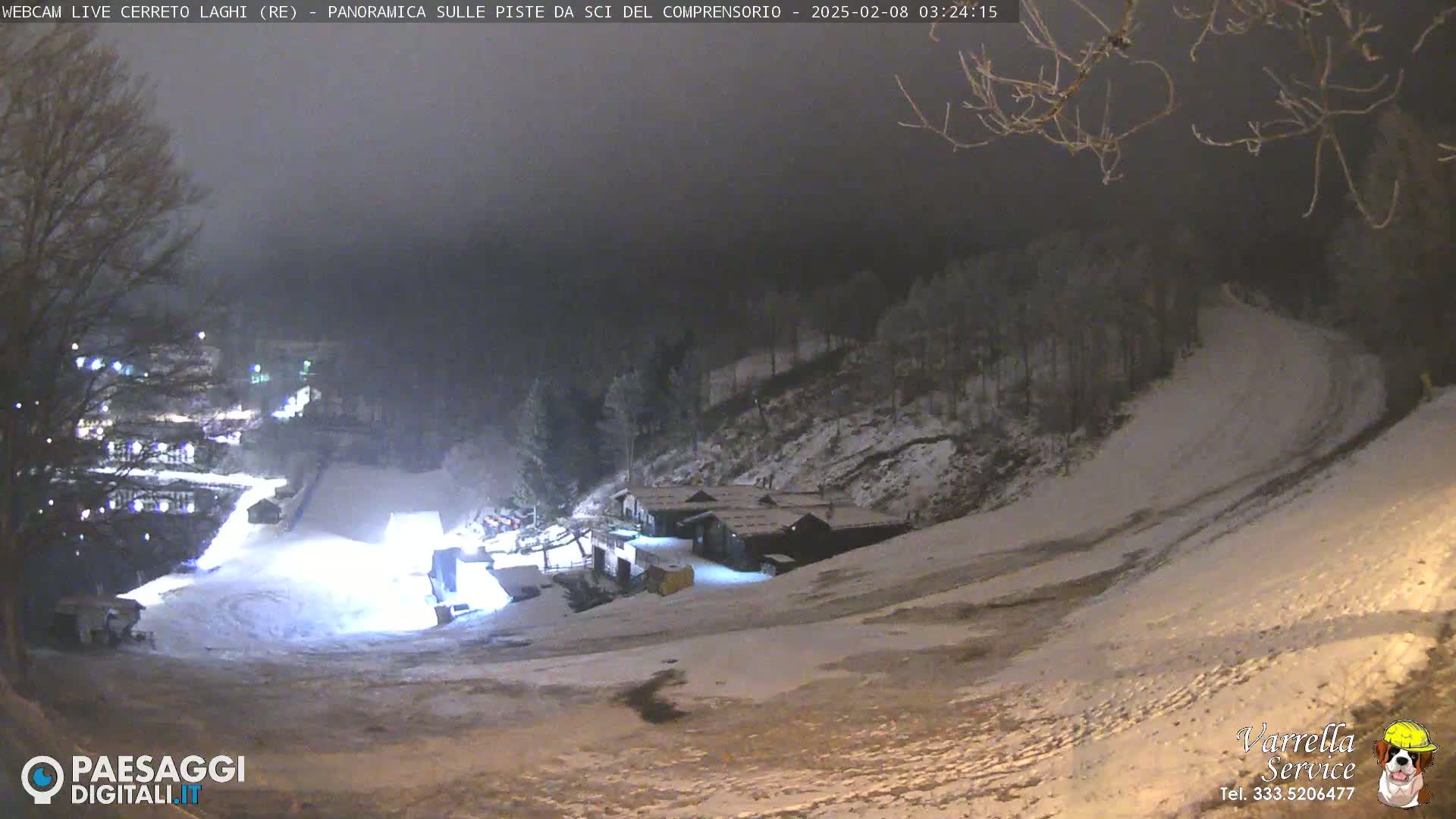 A nighttime view of a snow-covered ski slope with several buildings at the bottom, under a dark, overcast sky.