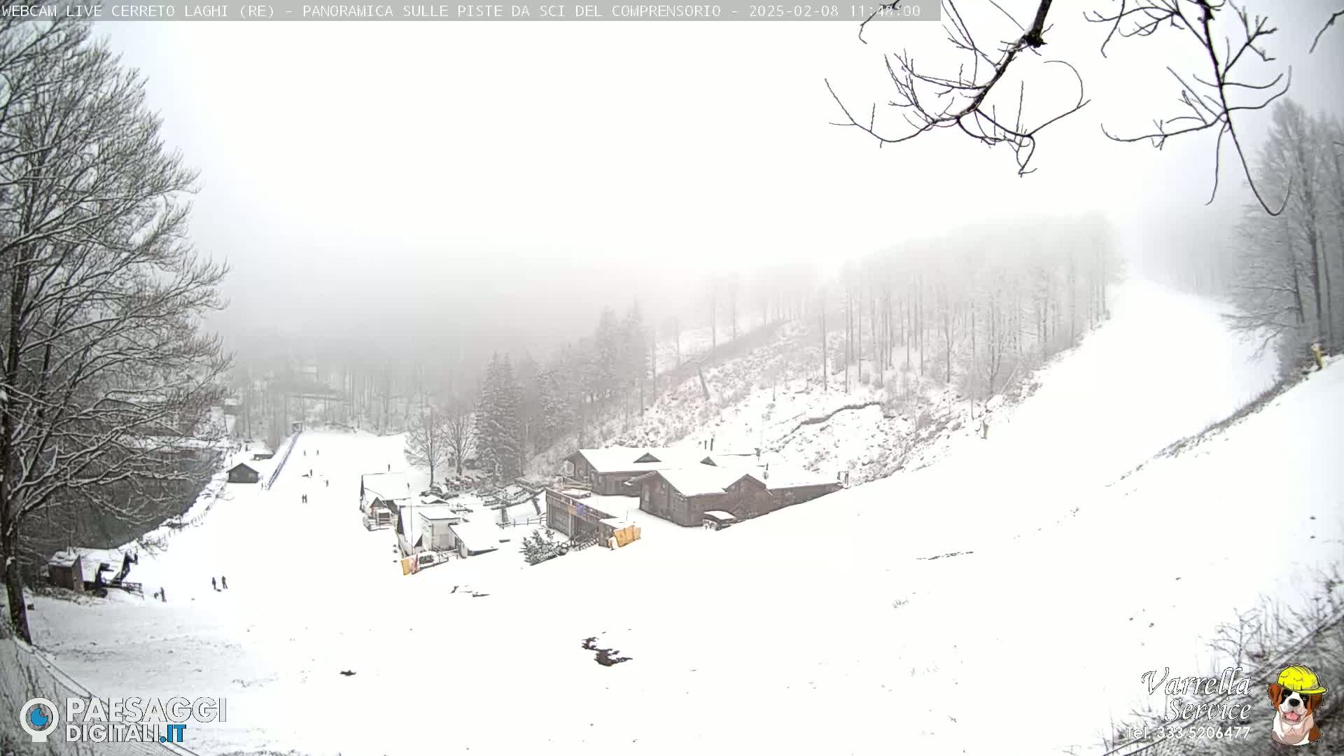 A snow-covered ski slope with a few skiers visible, several buildings, and bare trees in a foggy atmosphere.