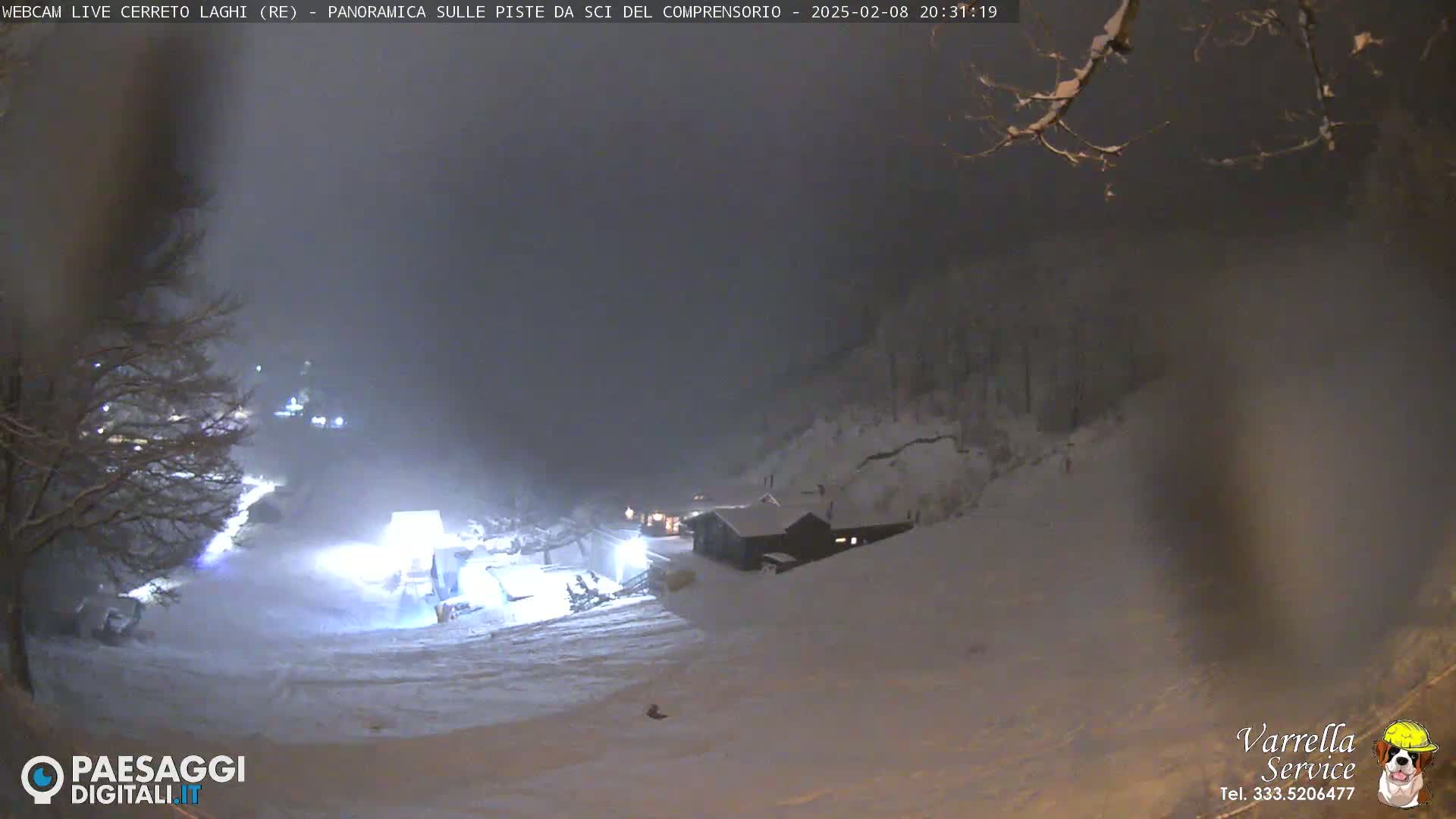 A snow-covered ski slope at night, with a small building and lights visible in the distance, under a dark, snowy sky.