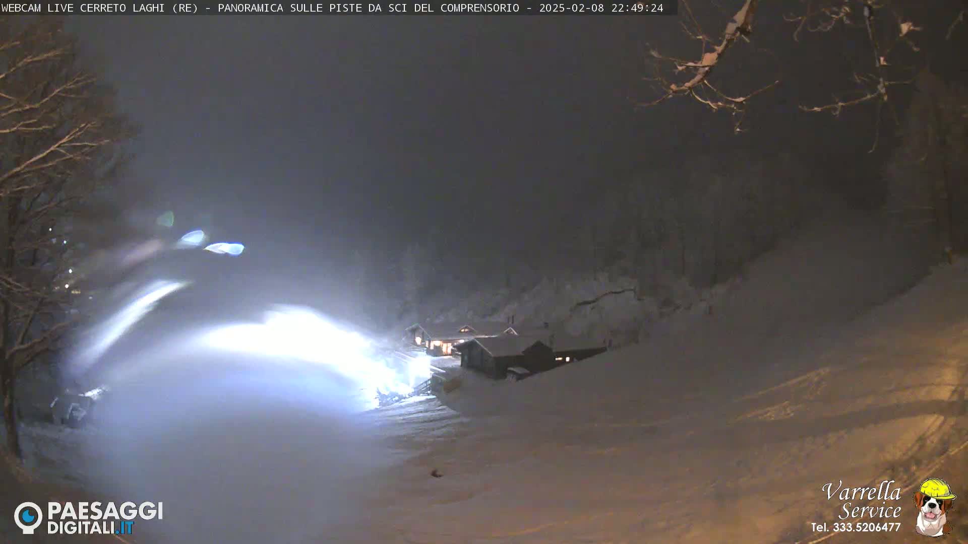 A snow groomer illuminates a snowy ski slope at night under a dark, overcast sky.