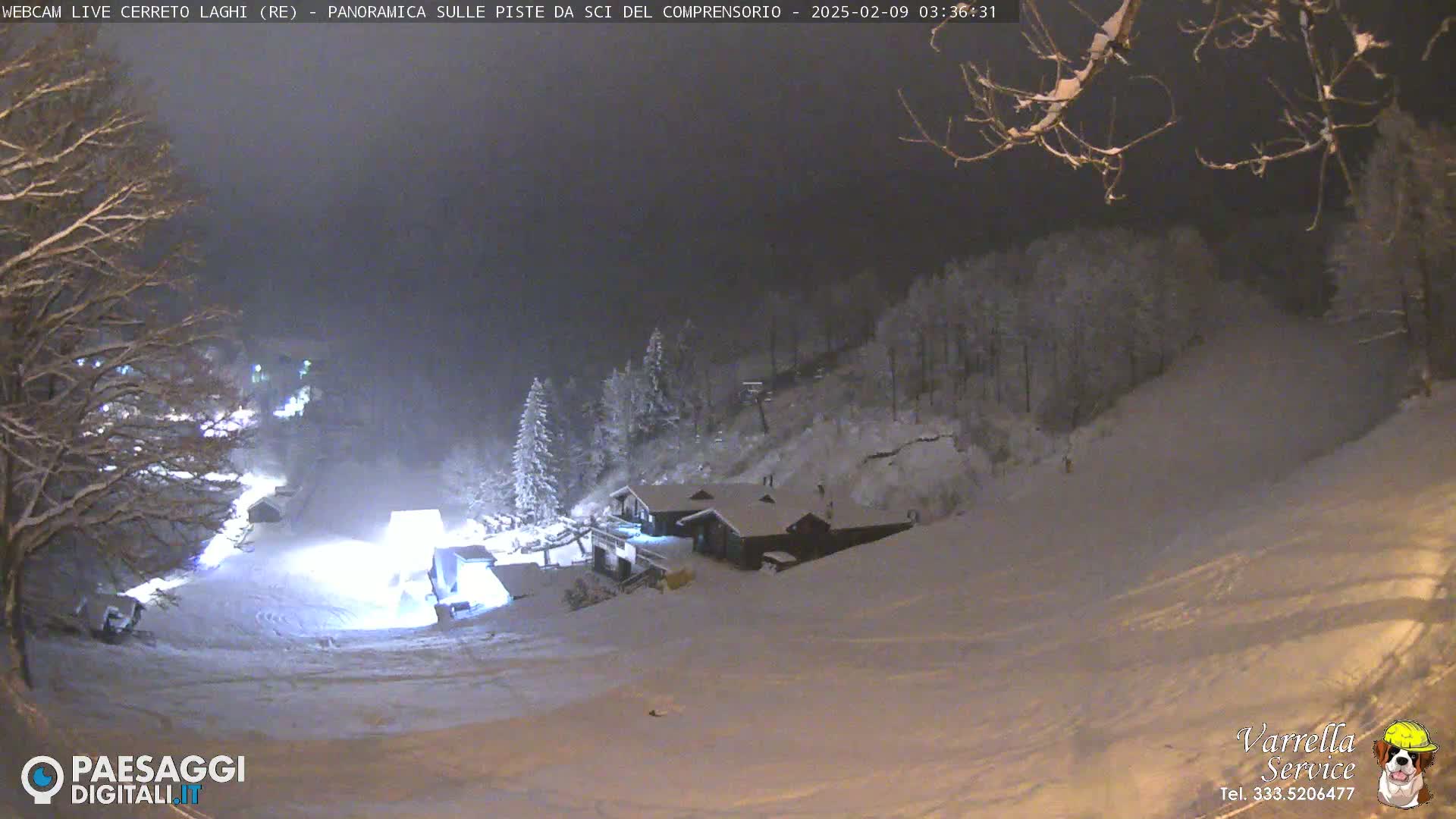 A snow-covered ski slope at night, illuminated by artificial lights, with a small building nestled amongst snow-covered trees.