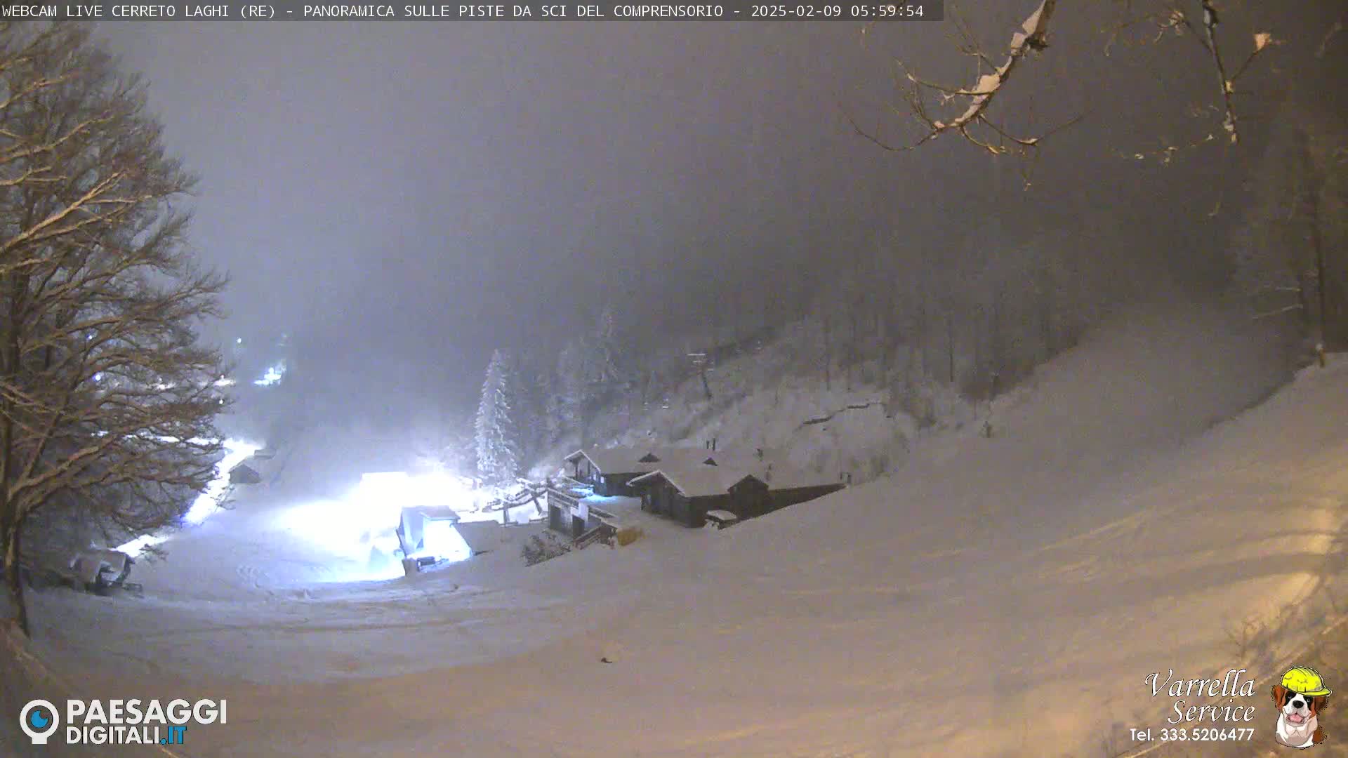 A snow-covered ski slope at night, illuminated by artificial lights, under a hazy, snowy sky.