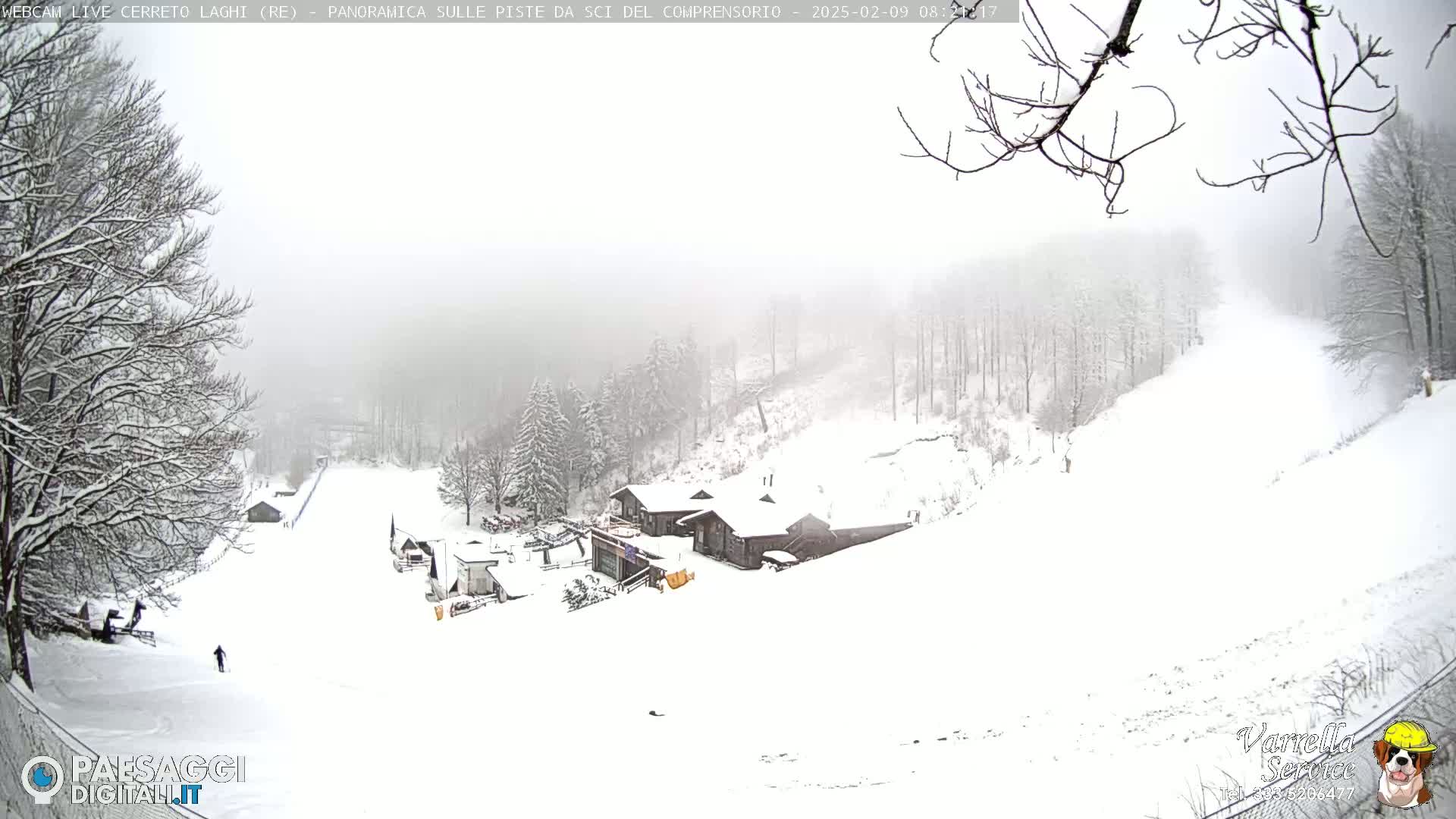 A snowy landscape shows a ski slope with a few buildings and snow-covered trees under an overcast sky.