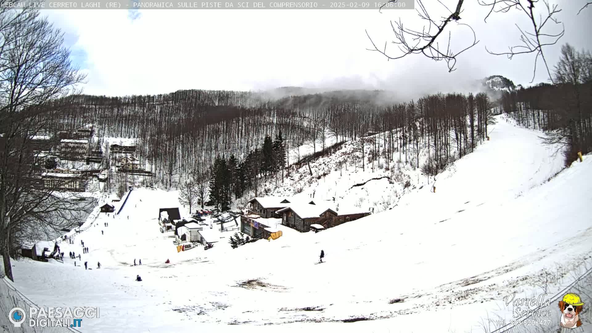 A snow-covered ski slope with a few people skiing and buildings in the background under a partly cloudy sky.