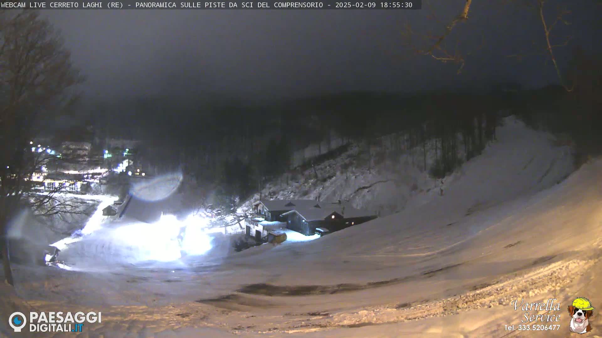 A nighttime, snow-covered ski slope is illuminated, showing a building and snow grooming equipment at the bottom of the hill under a dark sky.