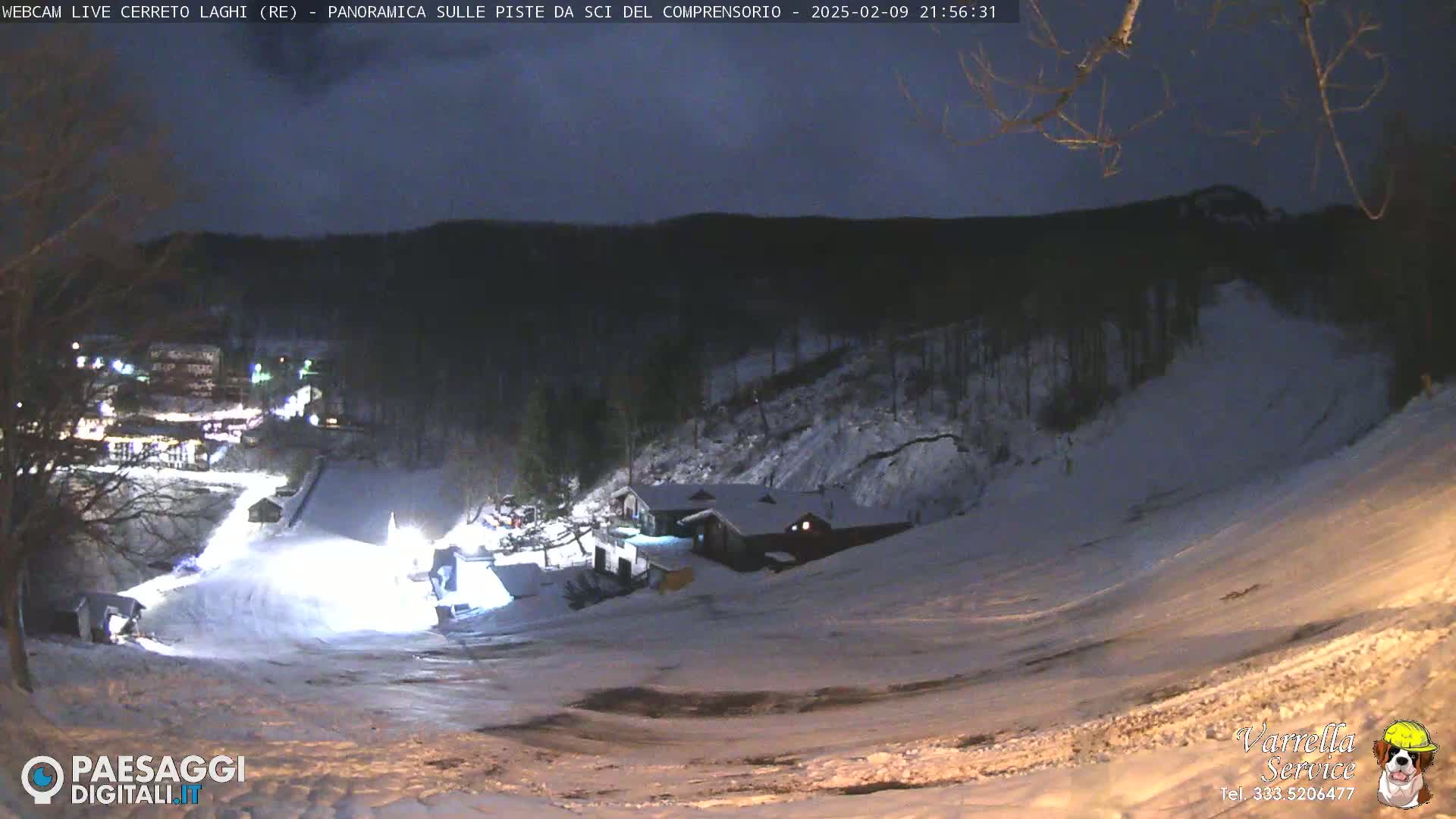 A snow-covered ski slope at night, illuminated by artificial lights, with a building and trees in the background under a dark sky.