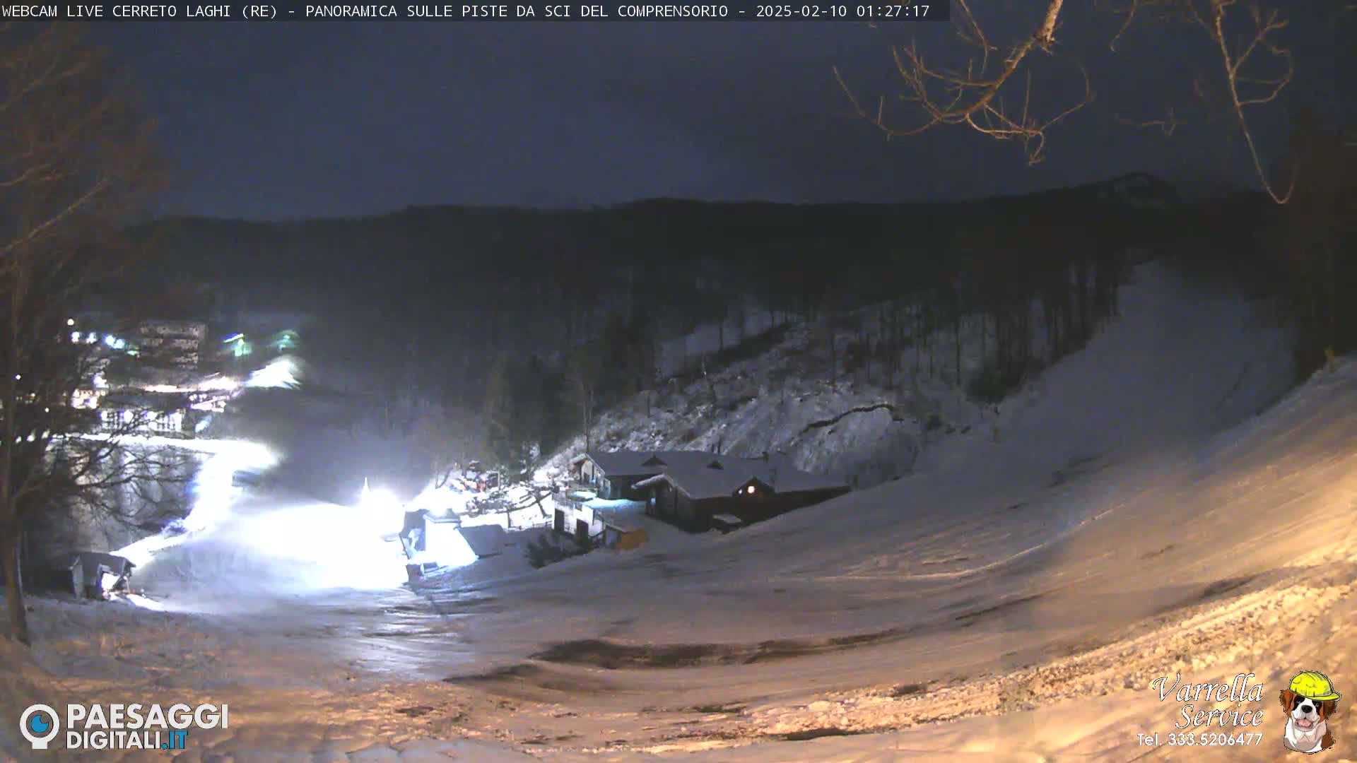 A snow-covered ski slope at night, illuminated by lights, with a building and trees in the background.