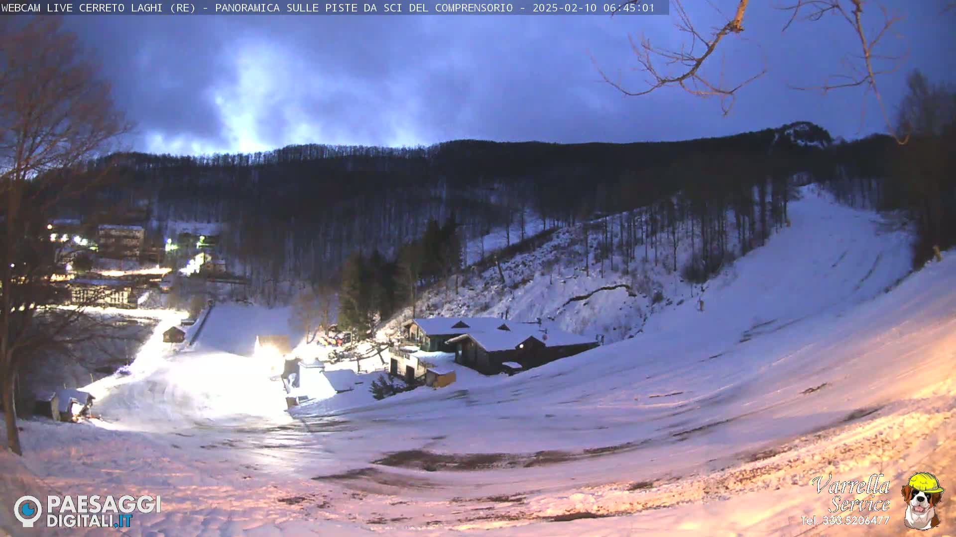 A snow-covered ski slope at twilight, illuminated by artificial lights, with a cluster of buildings near the bottom of the hill and a dark forest in the background.