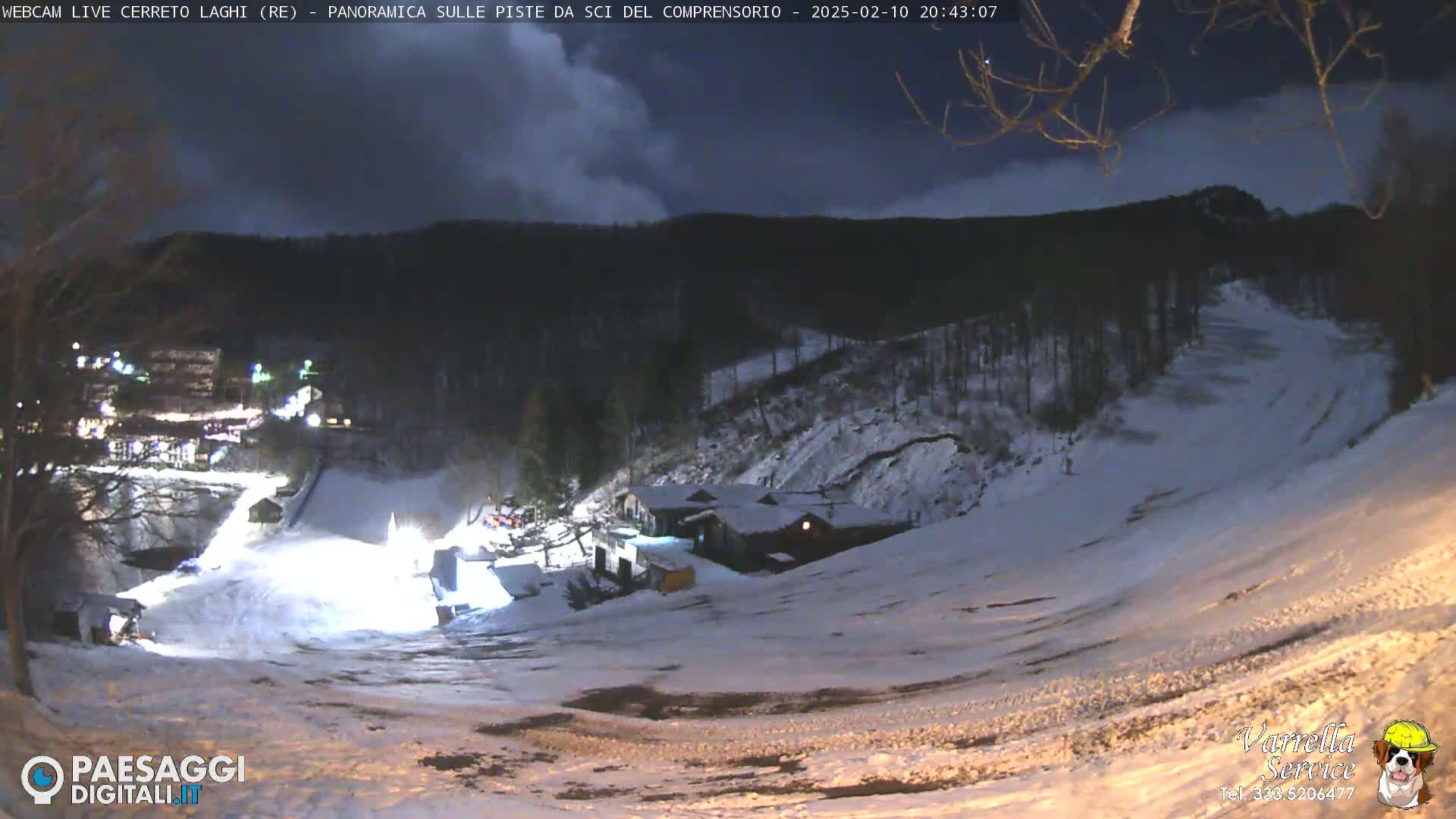 A nighttime view of a snow-covered ski slope with a building and lights visible in the distance under a cloudy sky.