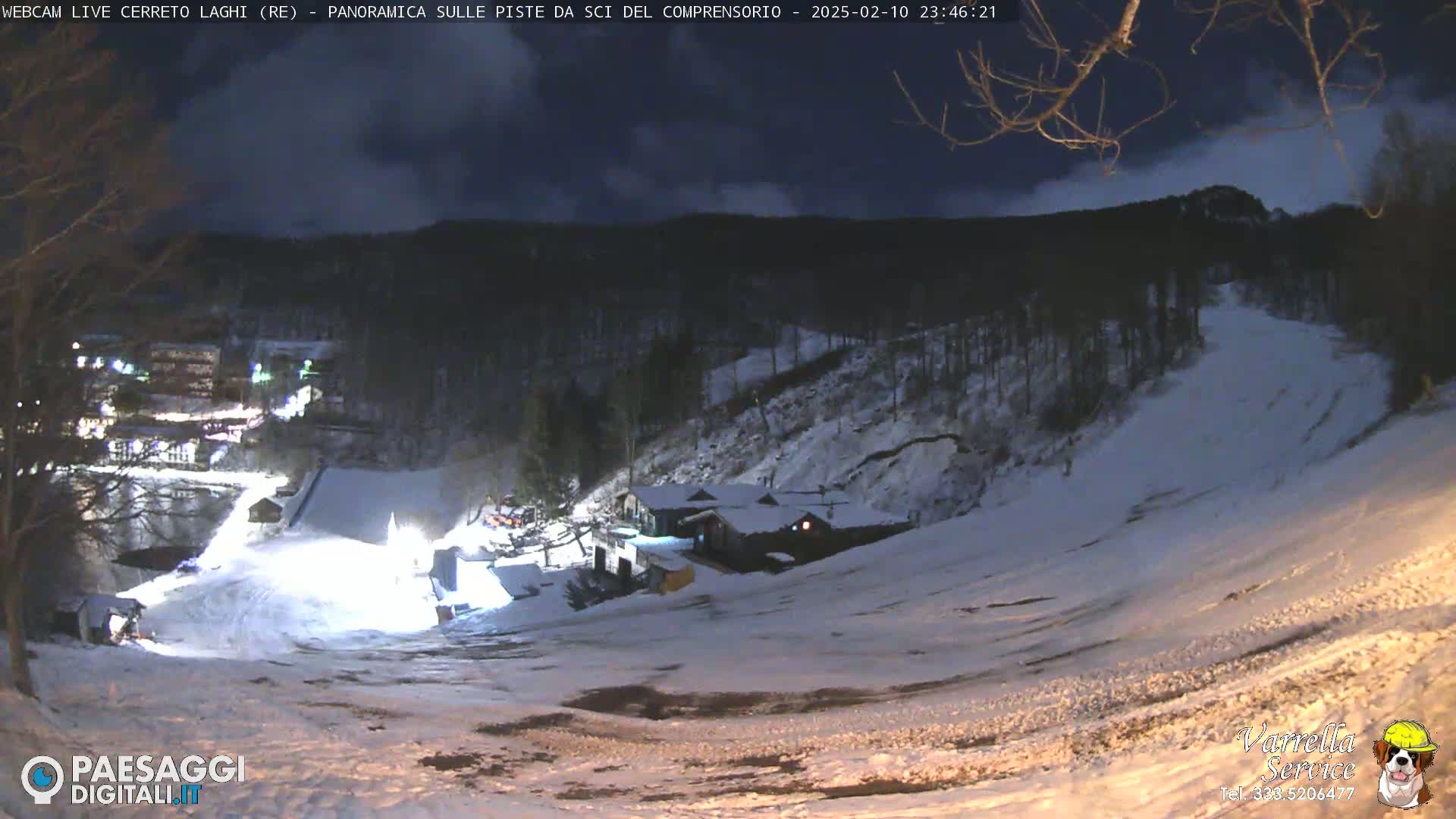 A nighttime, snow-covered ski slope is illuminated, with a small village visible in the distance under a partly cloudy sky.