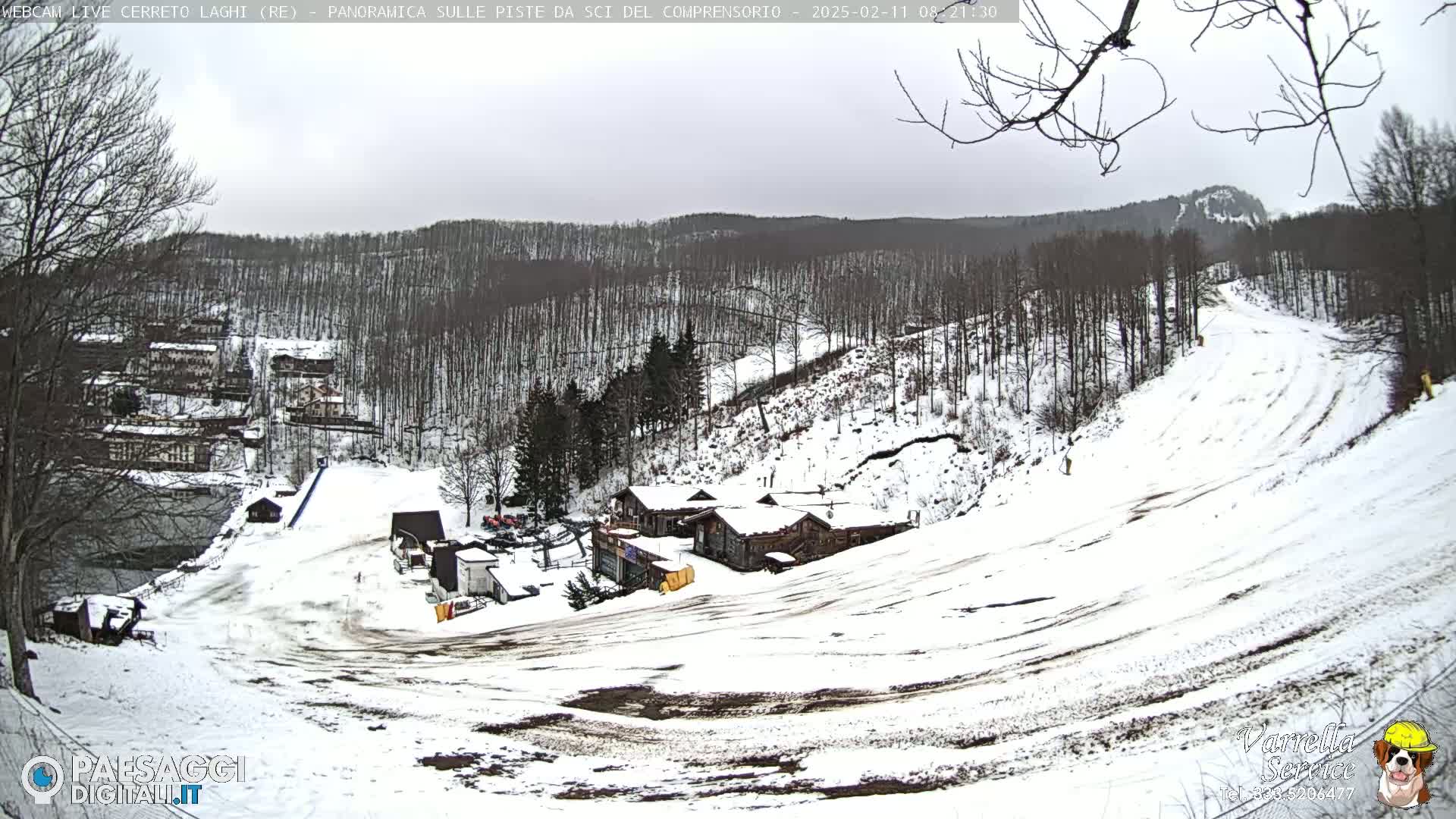 A snow-covered ski slope with several buildings and a wooded hillside in the background under an overcast sky.