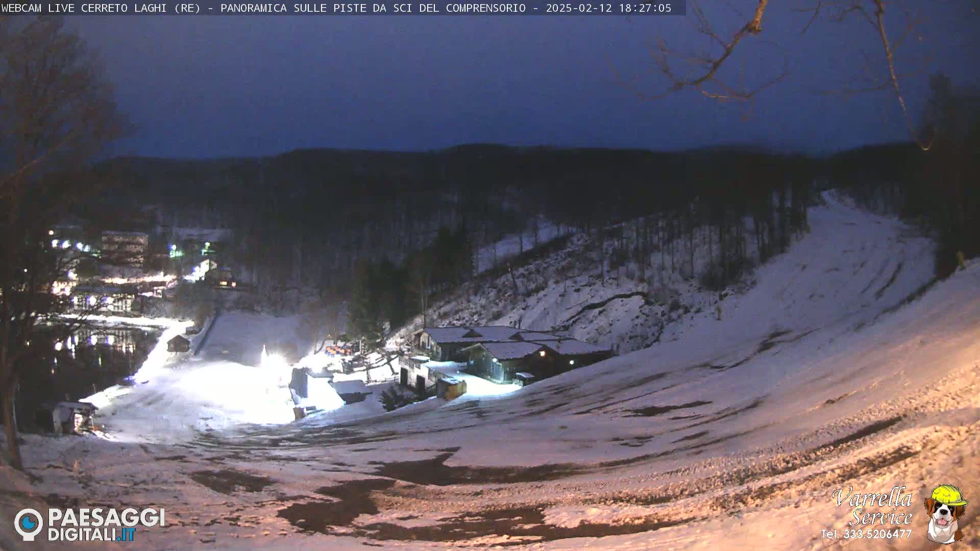 A snow-covered ski slope at night, illuminated by artificial lights, with a building at the bottom of the hill and a dark, wooded area in the background.