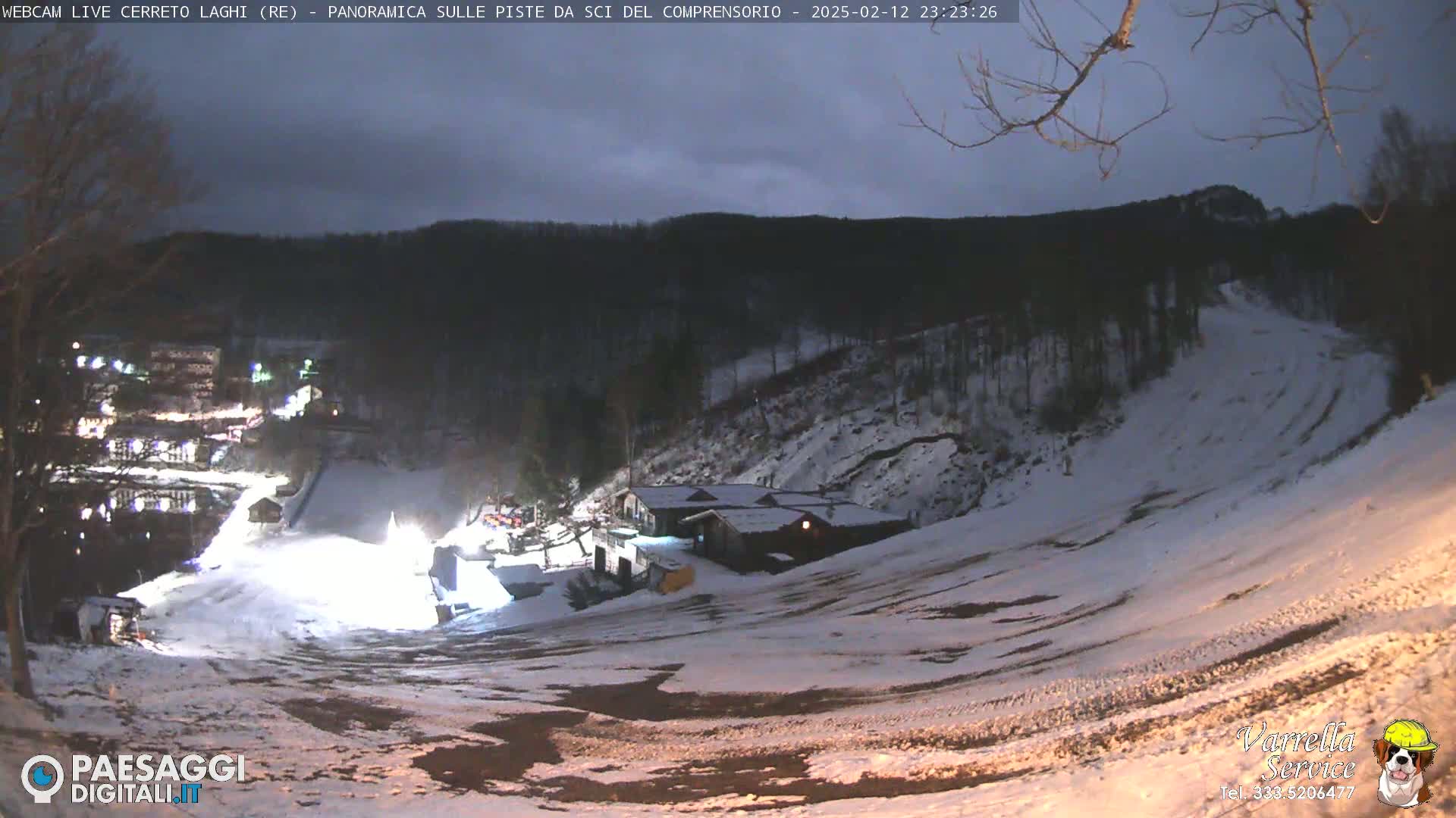 A nighttime, snow-covered ski slope with a building and lights visible under a partly cloudy sky.
