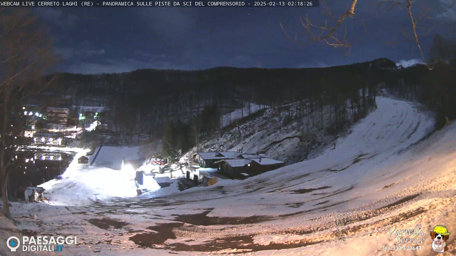 A snow-covered ski slope at night, illuminated by artificial lights, with a building and surrounding trees visible in the background under a dark sky.