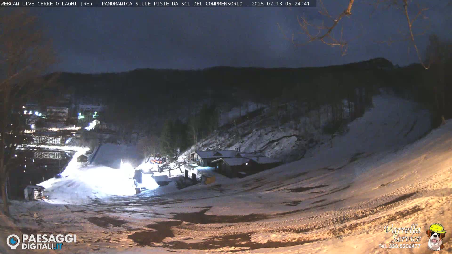 A snow-covered ski slope at night, illuminated by artificial lights, with a building and trees visible in the background under a dark sky.