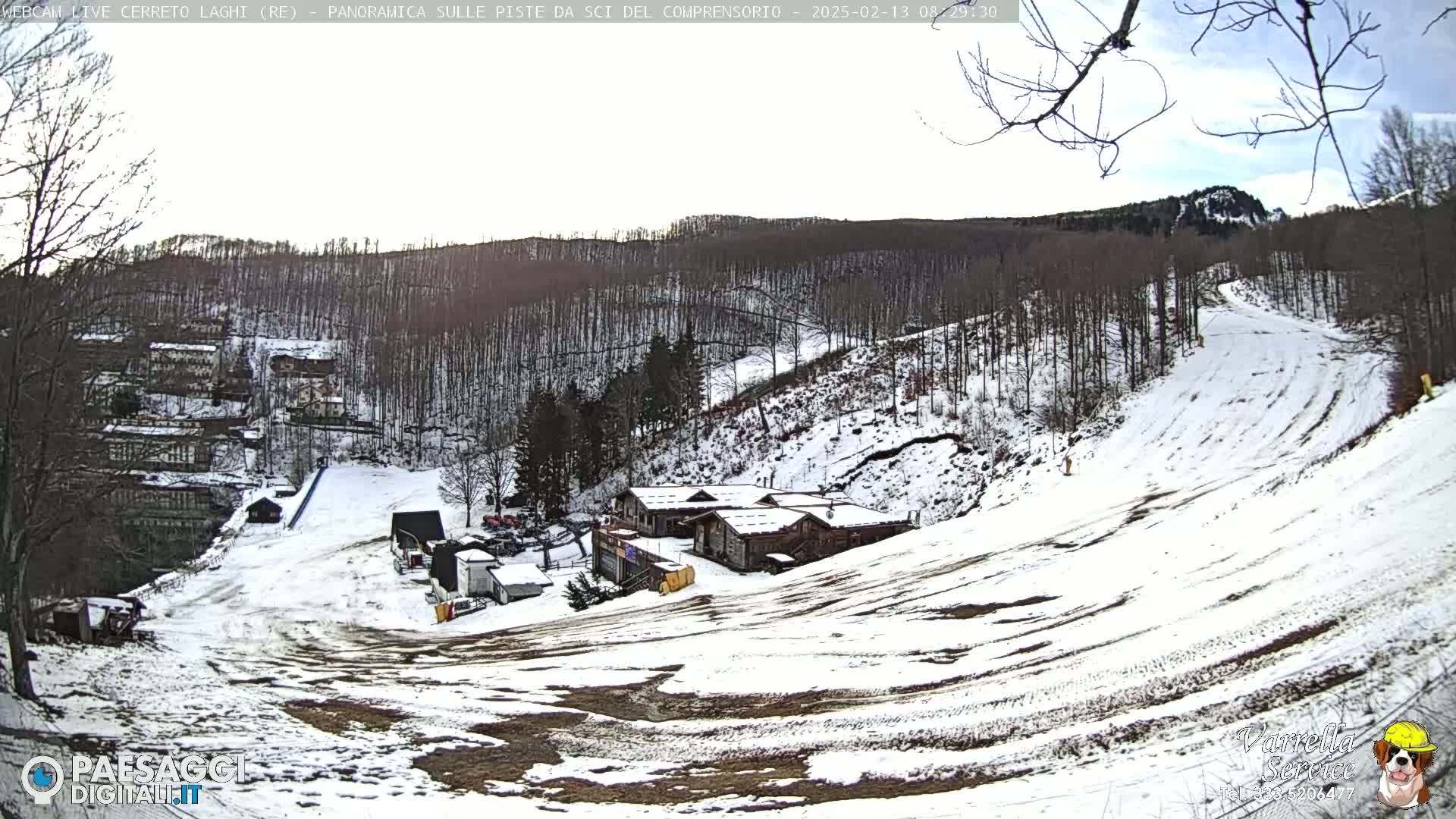 A snow-covered ski slope with a few buildings and a mostly leafless forest in the background under a partly cloudy sky.