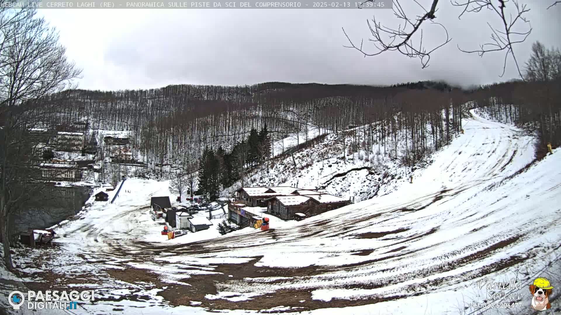 A snow-covered ski slope winds down a hill, past several buildings, toward a wooded area under an overcast sky.
