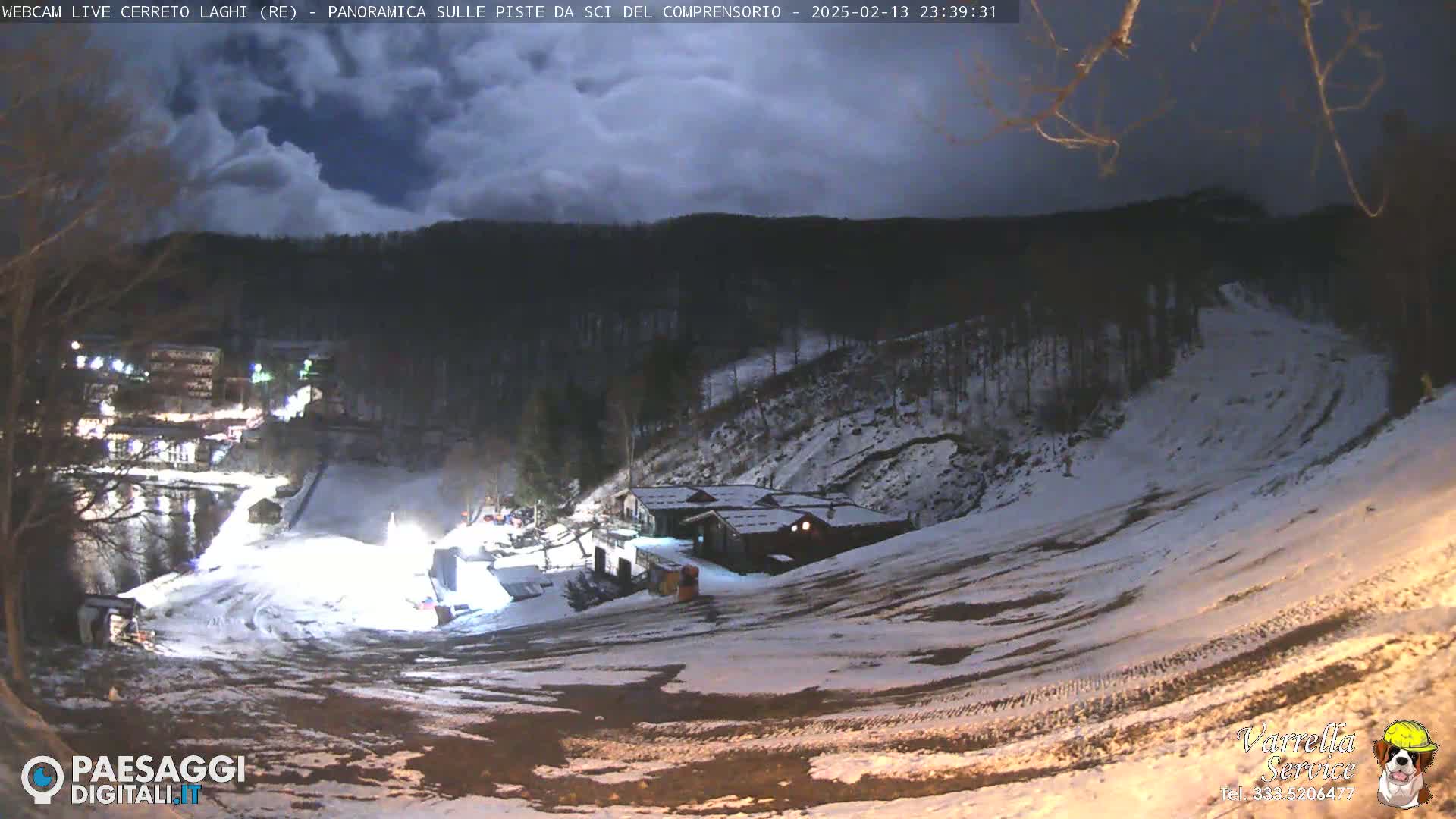 A nighttime view of a snow-covered ski slope with a few buildings and lights visible under a partly cloudy sky.