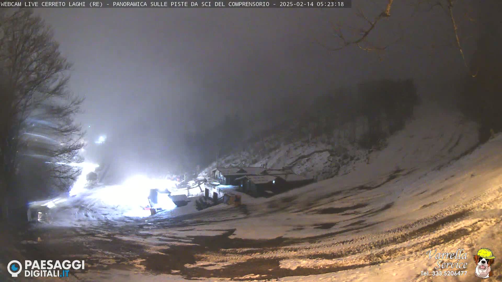 A snow-covered ski slope at night under foggy conditions, with a building and lights visible in the distance.