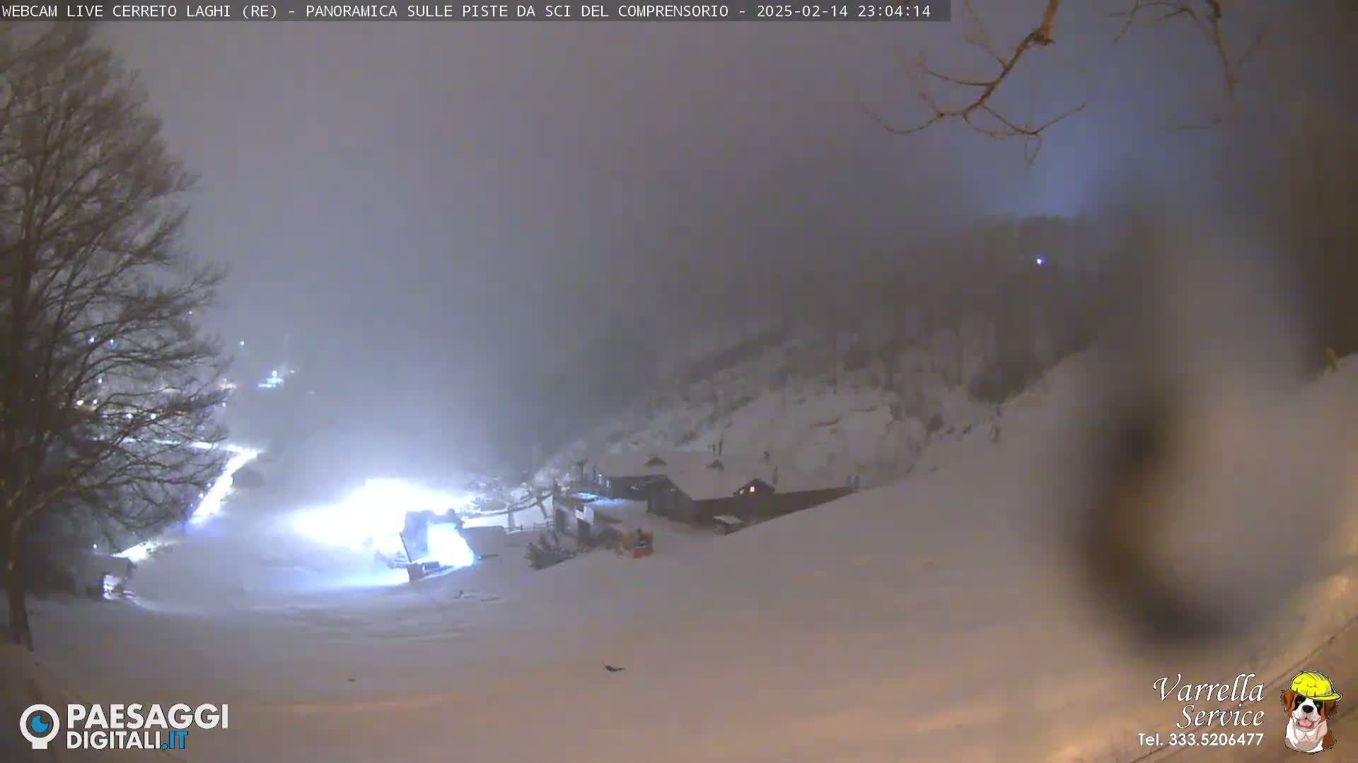 A snow-covered ski slope at night is dimly lit, with a building and trees visible through a hazy atmosphere.