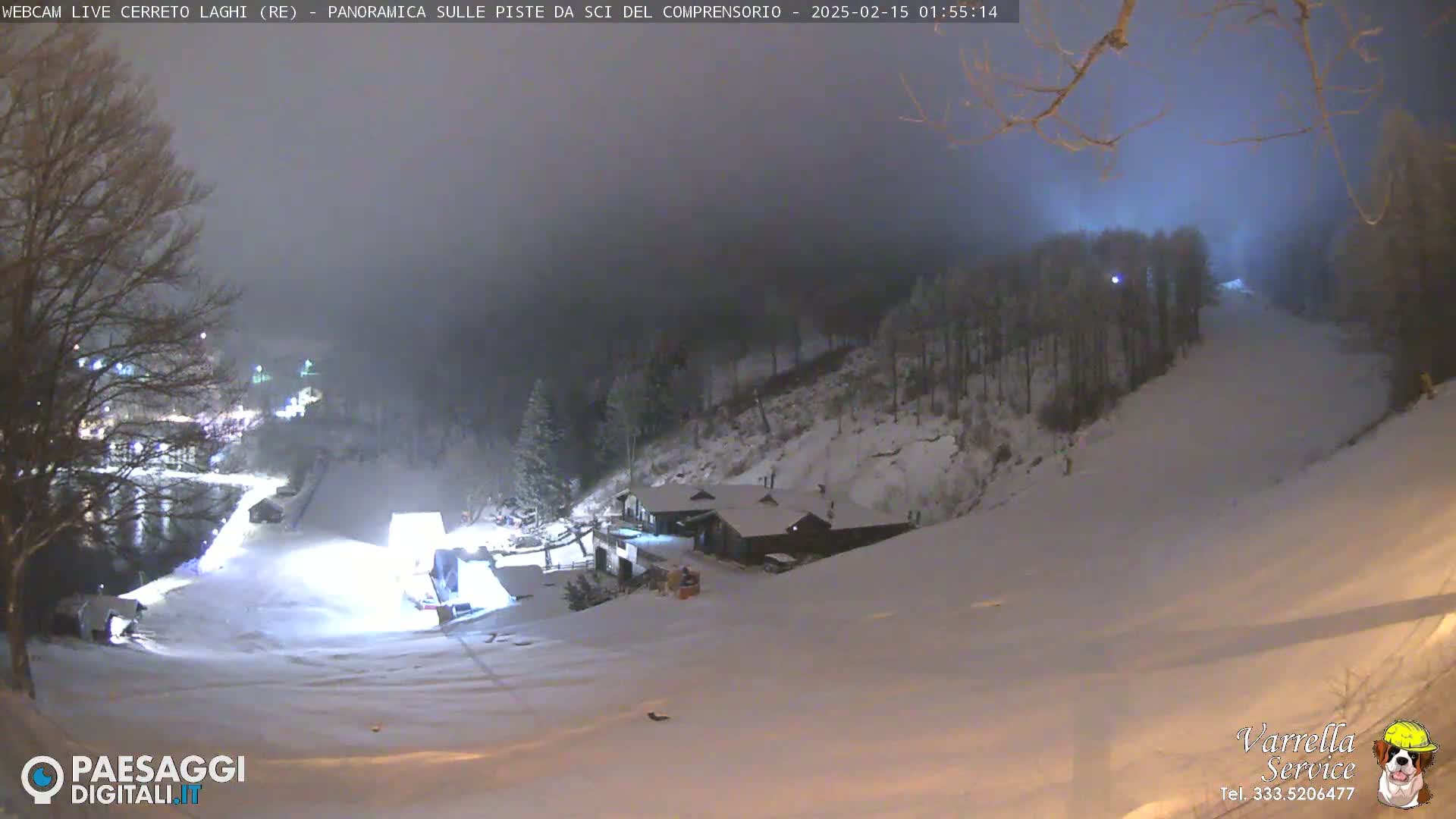 A snow-covered ski slope at night under a hazy sky, illuminated by artificial lights, with a building at the bottom of the slope.