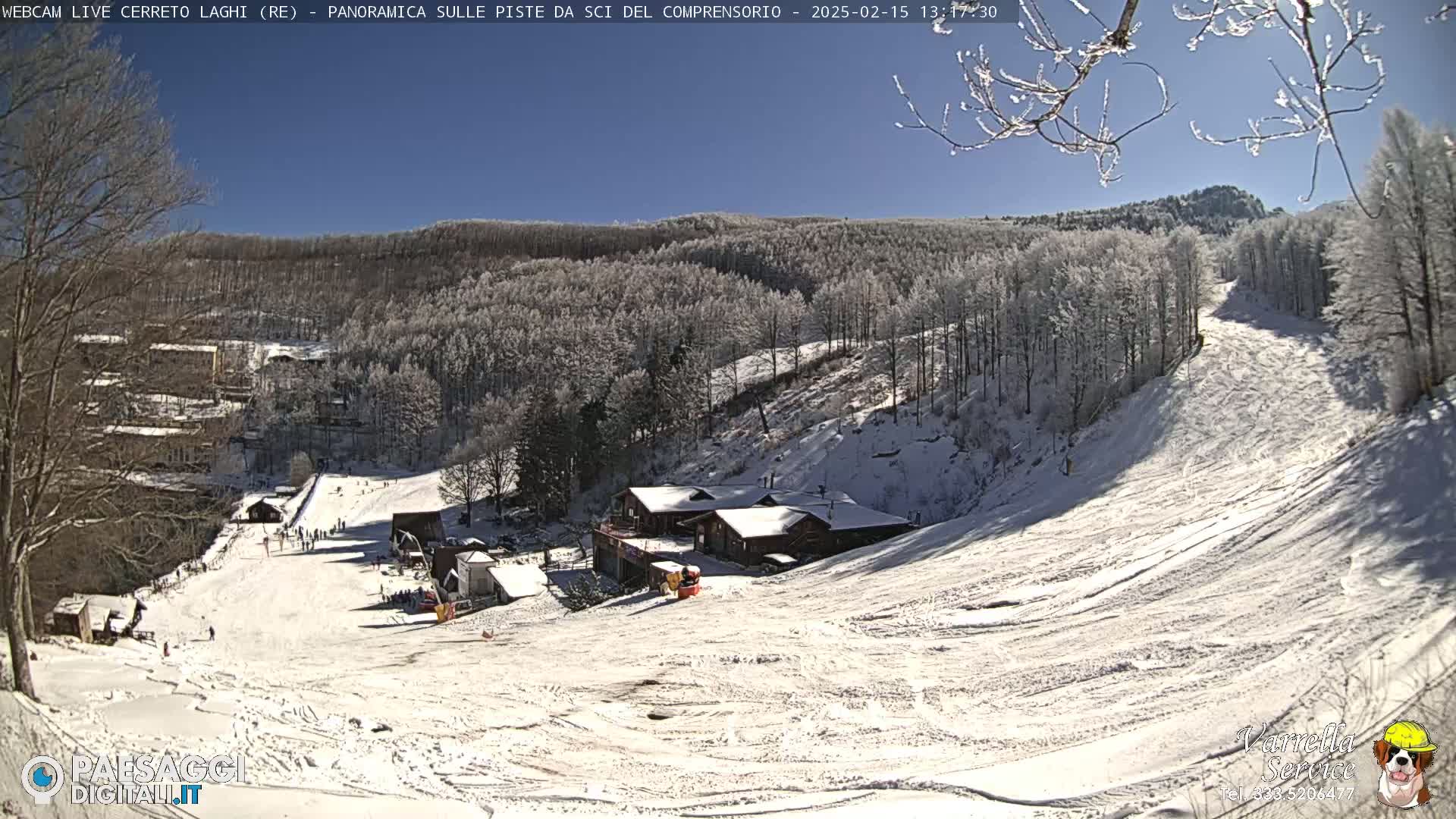 A sunny, snow-covered ski slope with several buildings and skiers visible, surrounded by snow-dusted trees on a hillside.