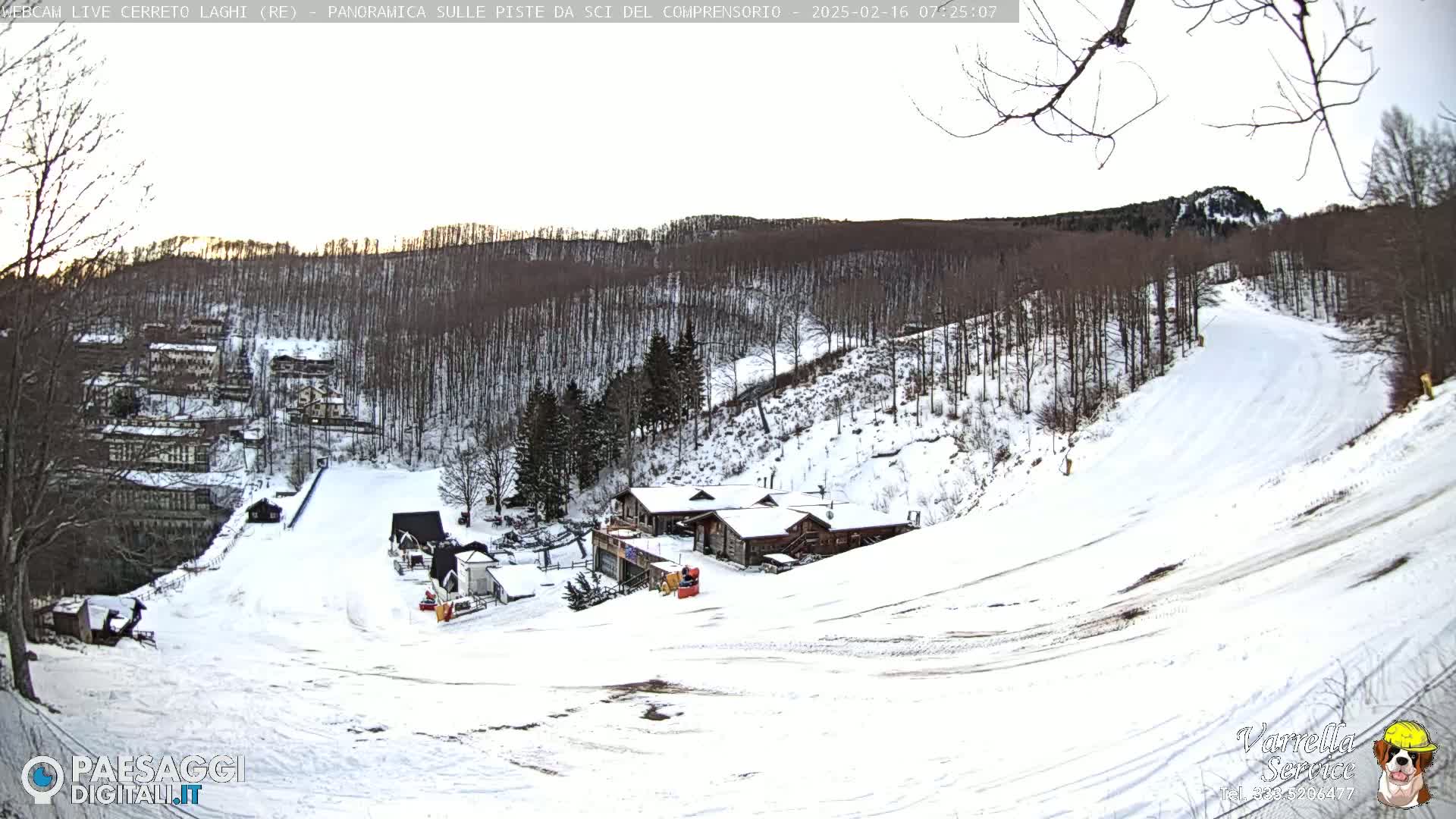 A snow-covered ski slope with several buildings and a forest in the background under a partly cloudy sky.