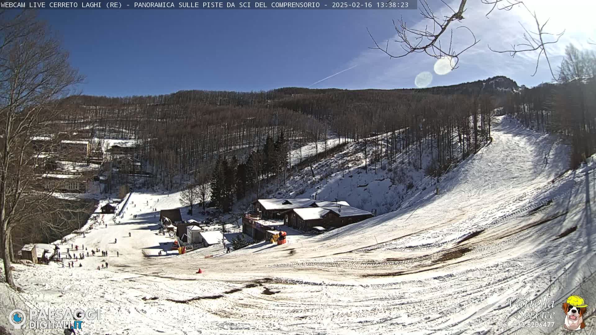 A sunny, snow-covered ski slope with a cluster of buildings at its base, surrounded by a snow-dusted forest under a clear blue sky.