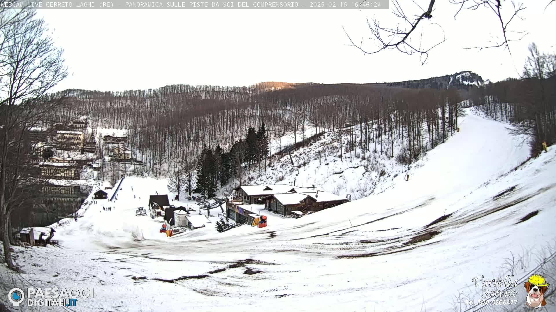 A snow-covered ski slope with several buildings and sparsely populated trees under a bright, mostly clear sky.