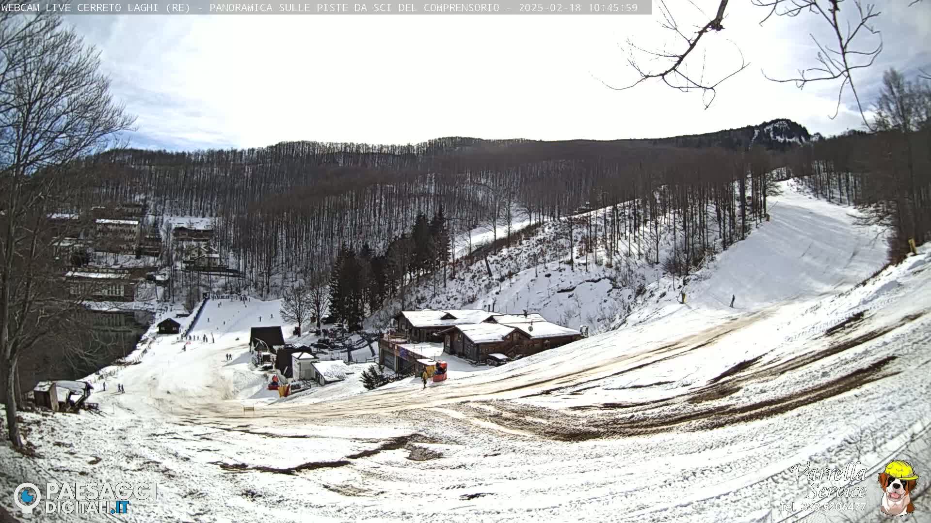 A snow-covered ski slope descends from a cluster of buildings nestled among snow-dusted trees under a mostly clear sky.