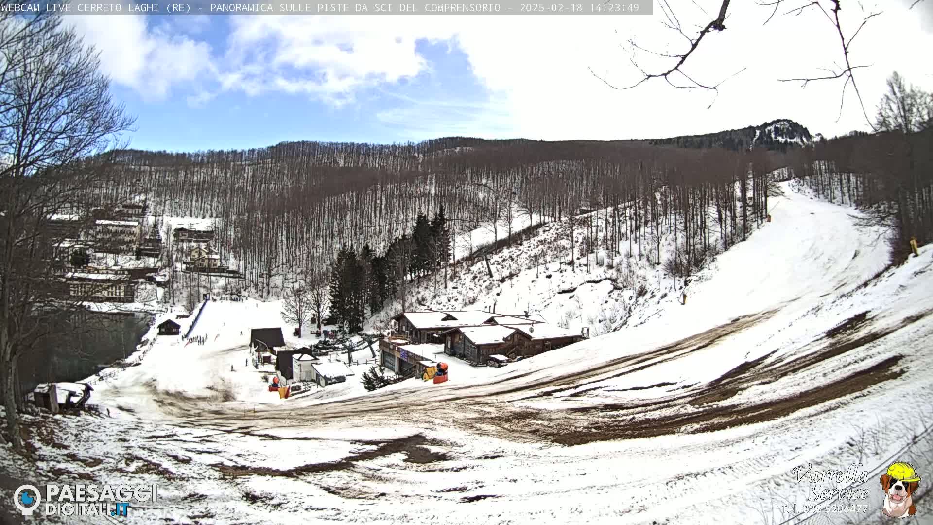 A snow-covered ski slope with a few buildings and a sparsely populated forest under a partly cloudy sky.
