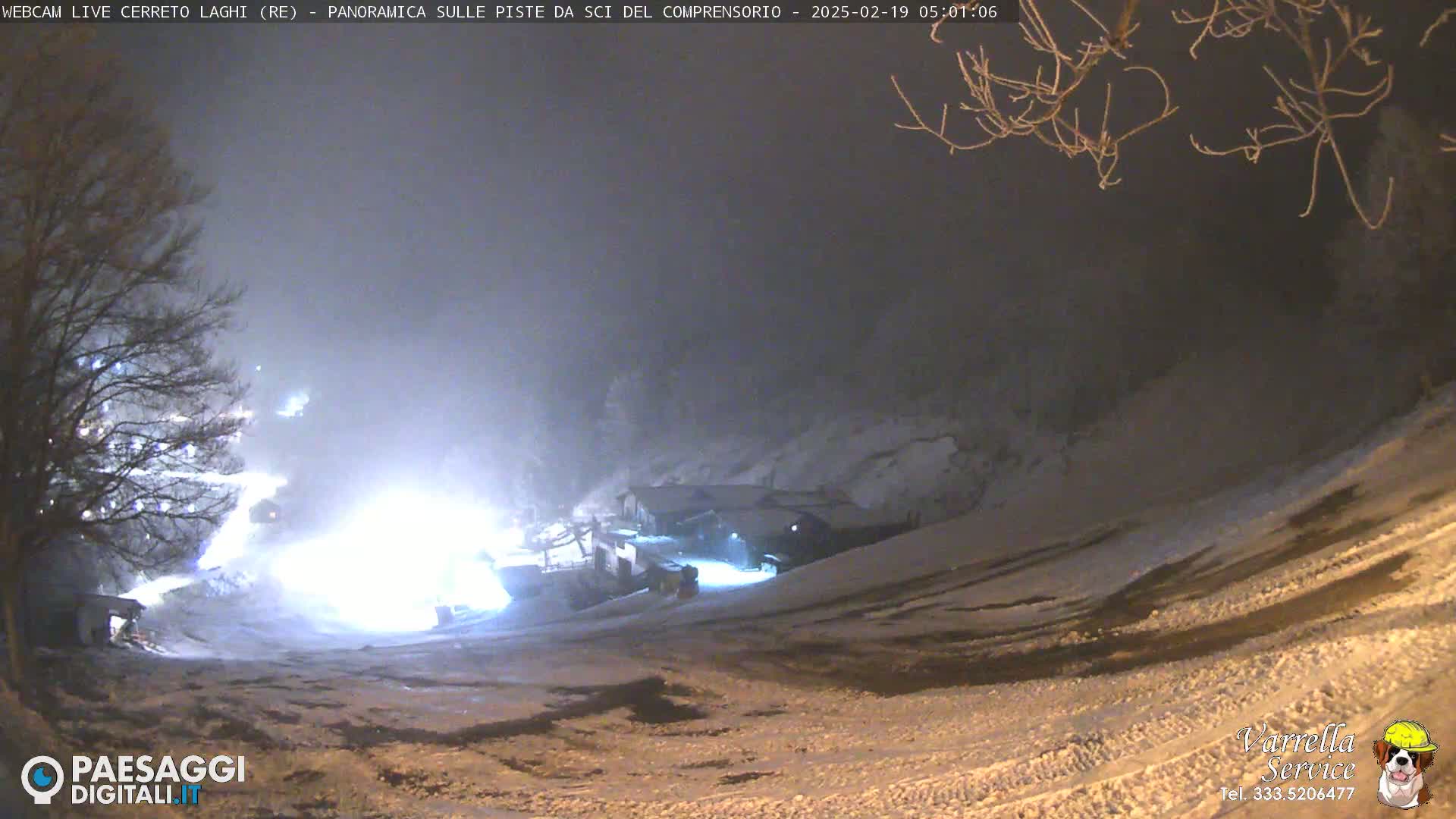 A nighttime, snowy scene shows a snow-covered slope with lights illuminating snow groomers near a building under a hazy sky.