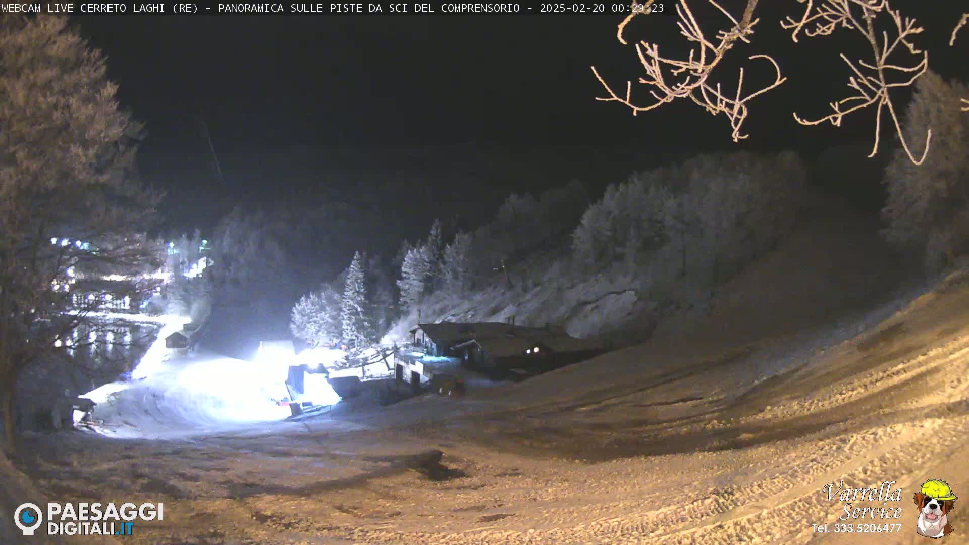 A snow-covered ski slope at night, illuminated by artificial lights, with a building and trees in the background.