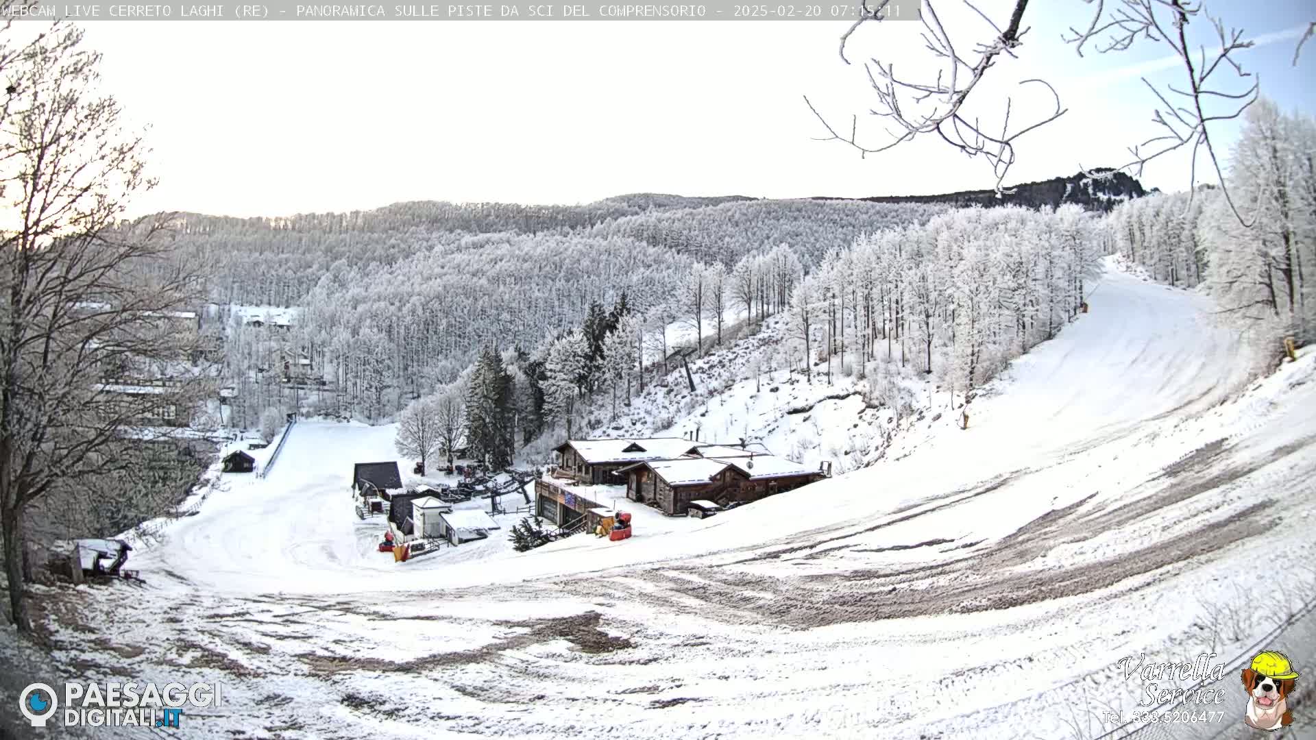 A snow-covered ski slope with several buildings at the bottom of the hill, surrounded by snow-covered trees under a mostly clear sky.