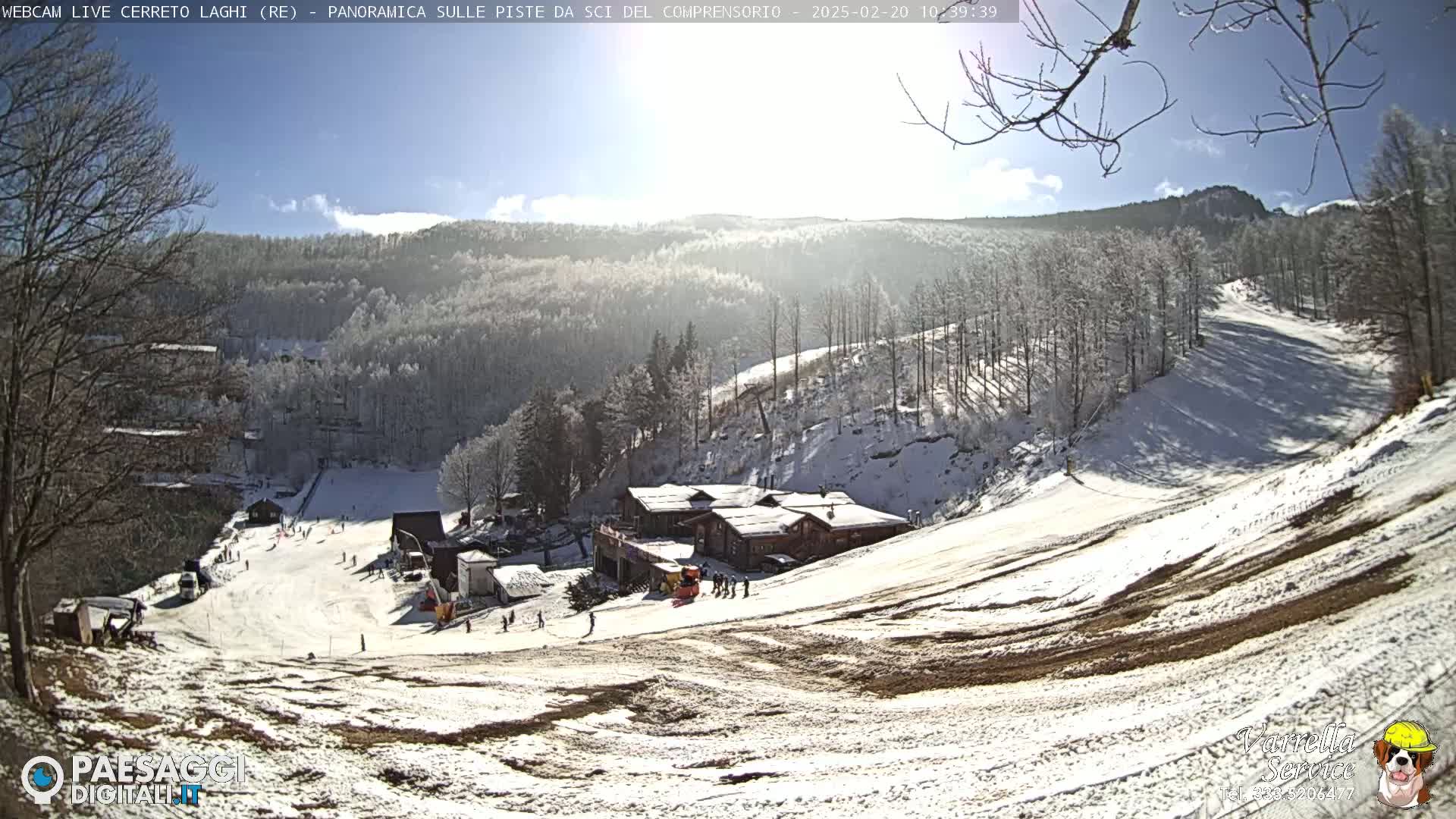 A sunny winter day at a snow-covered ski resort shows a downhill slope with several skiers and a cluster of buildings.