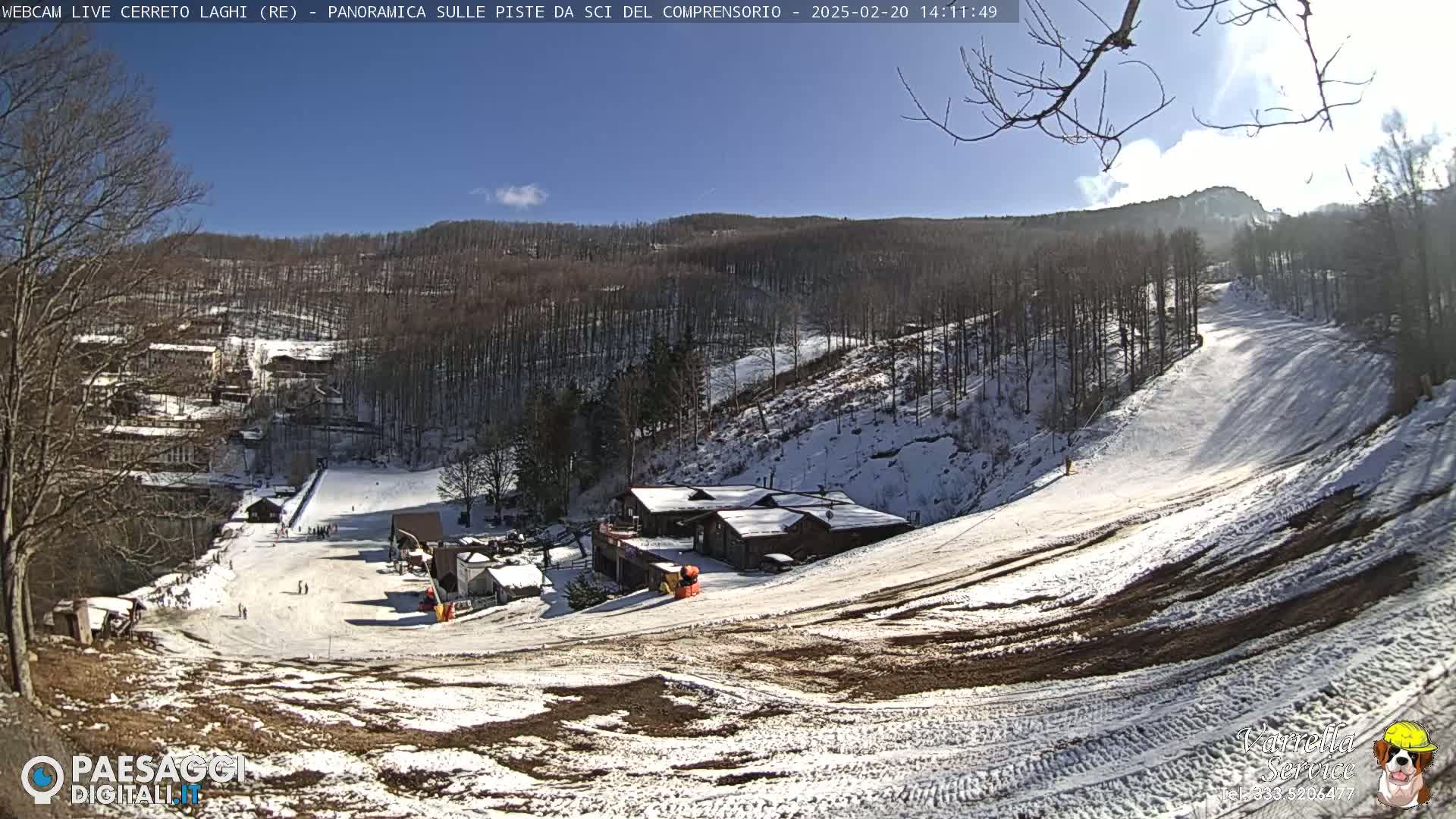 A snow-covered ski slope with a few skiers, several buildings, and a mostly sunny sky.